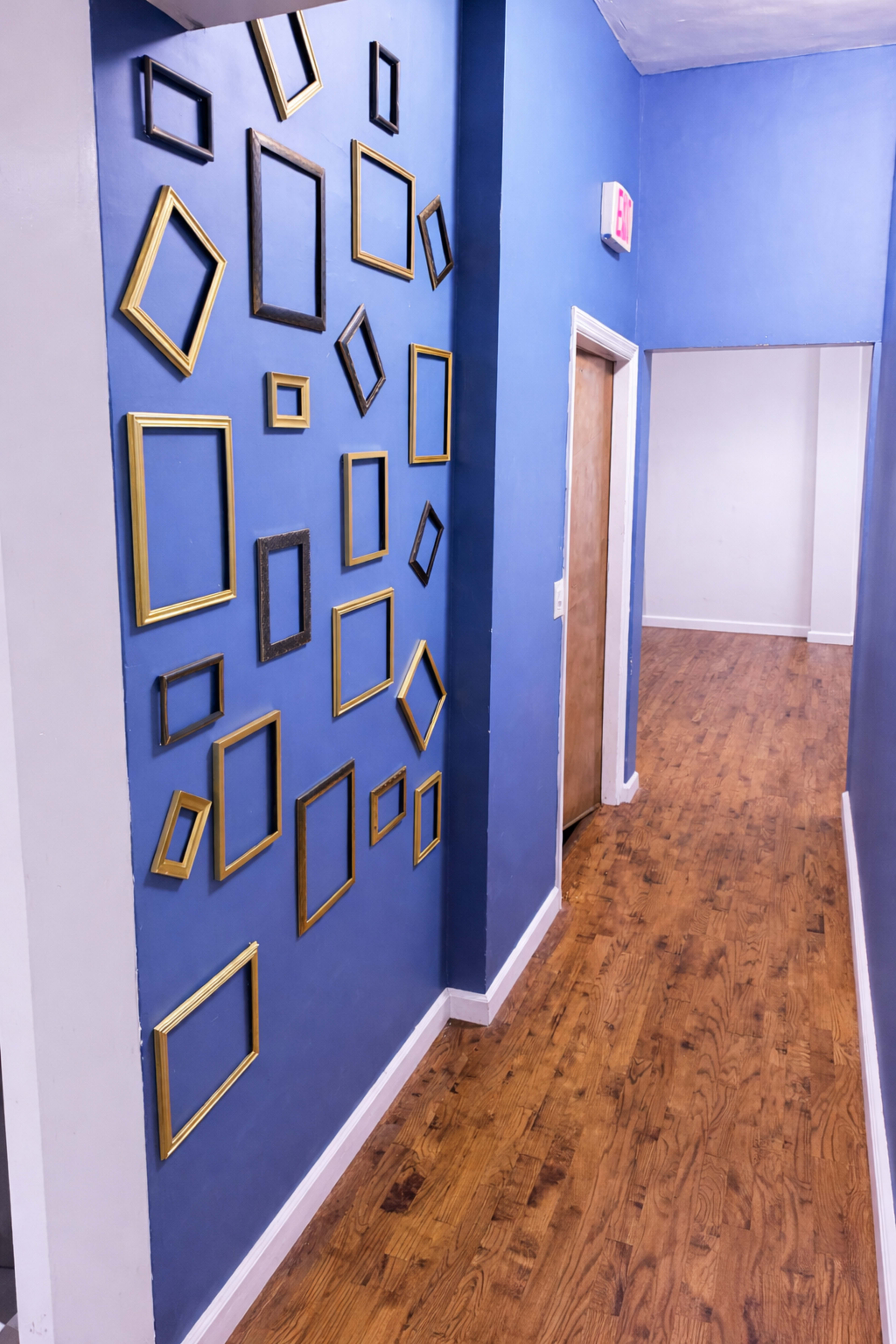 The image shows a hallway with a blue wall adorned with multiple empty picture frames of various sizes and shapes, leading to a wooden door at the end.