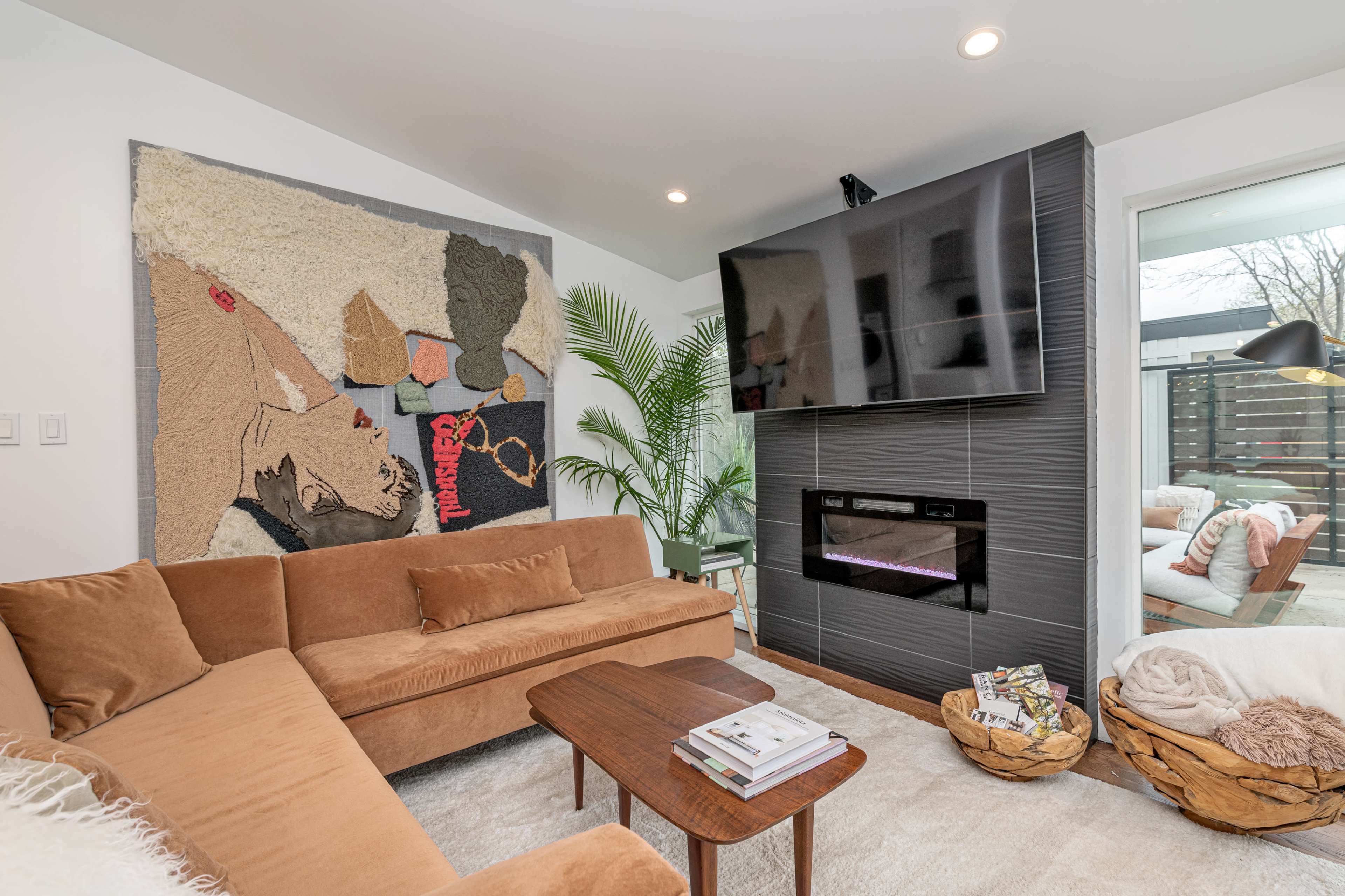 A modern living room featuring a brown sectional sofa, a wall-mounted television, and a decorative tapestry above a sleek electric fireplace.