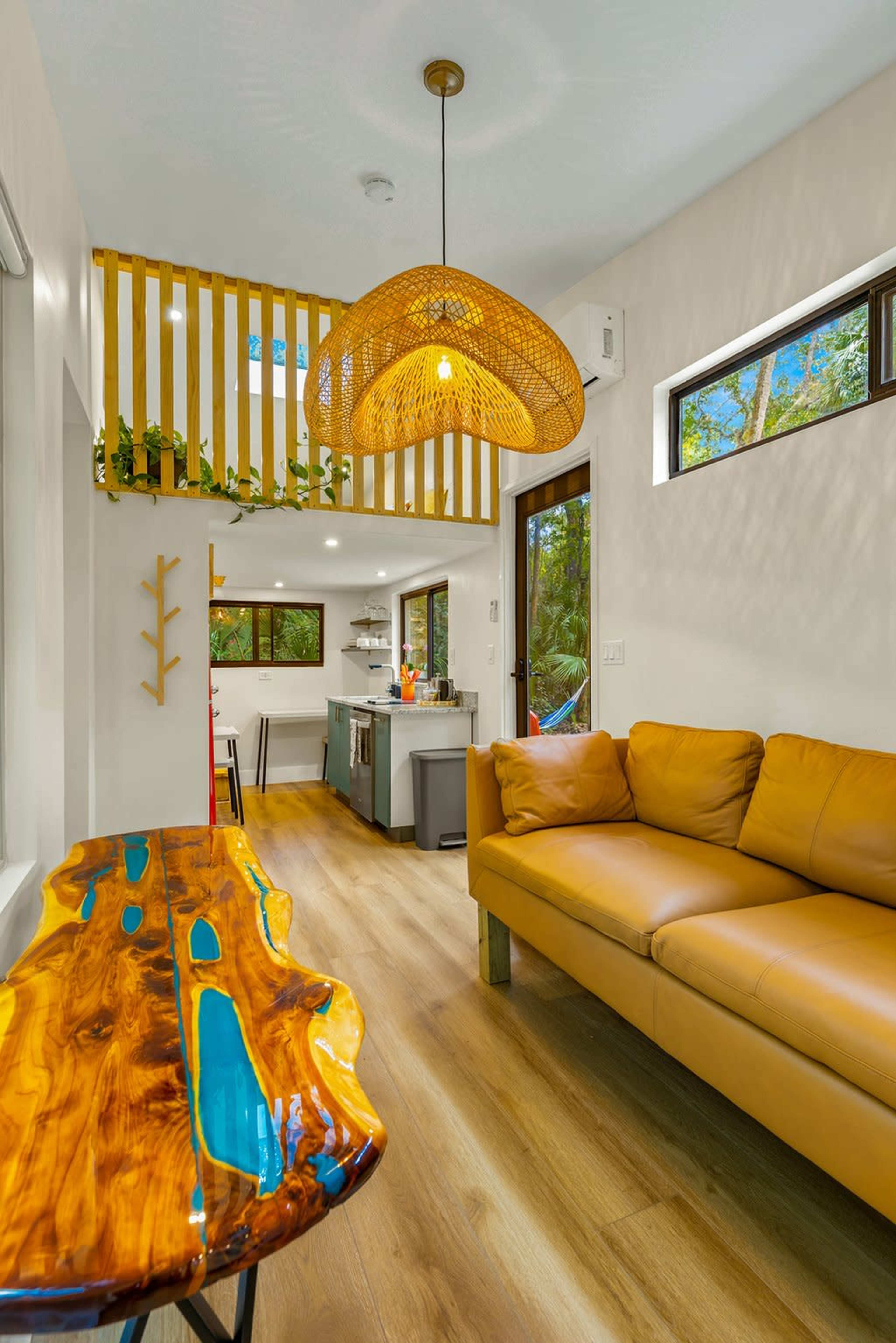 The image shows a modern interior of a living space featuring a wooden table, a tan leather sofa, and a kitchen area in the background.