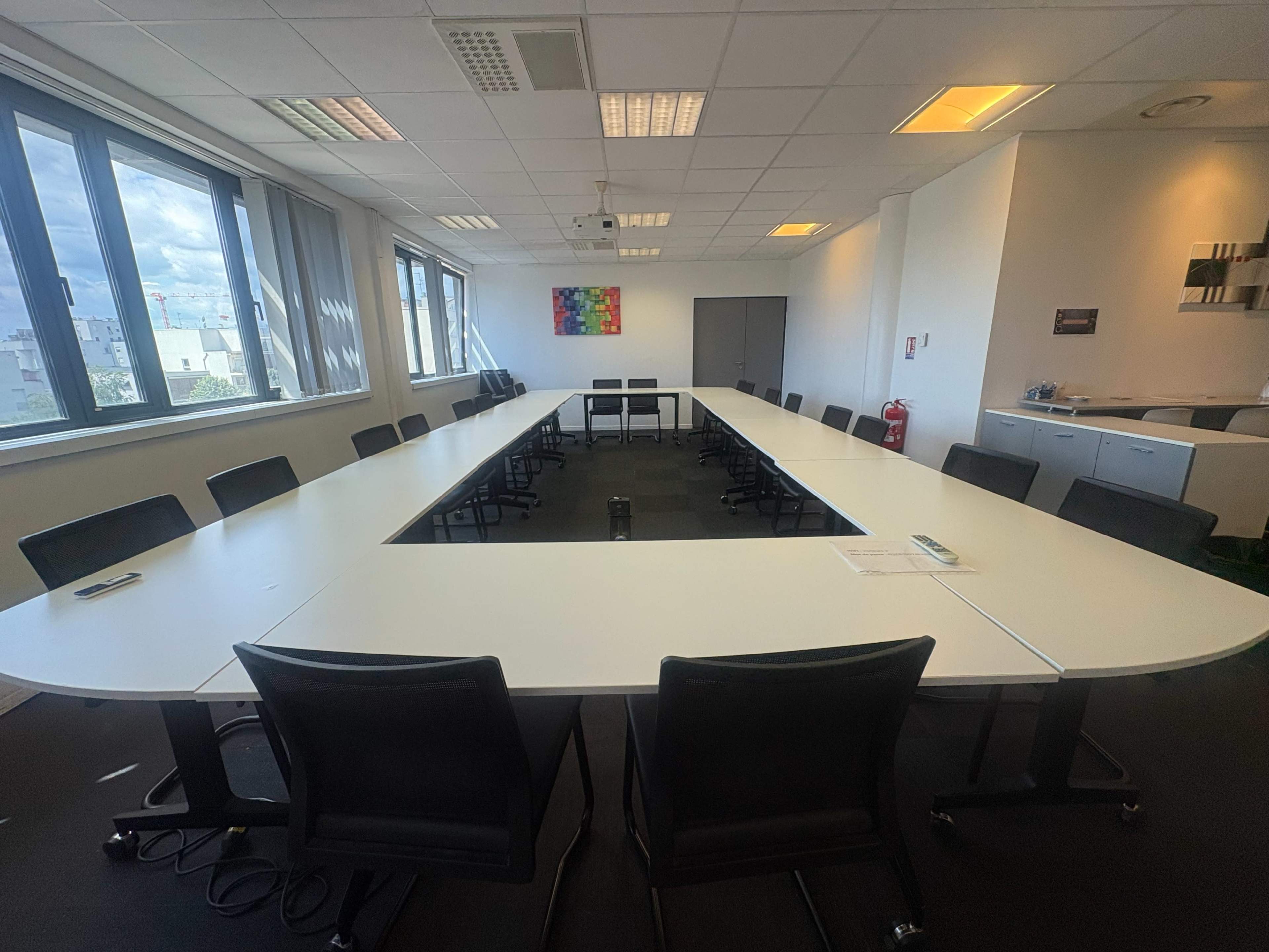 A large, rectangular conference room features a white table surrounded by black chairs, with windows providing natural light and a modern design aesthetic.
