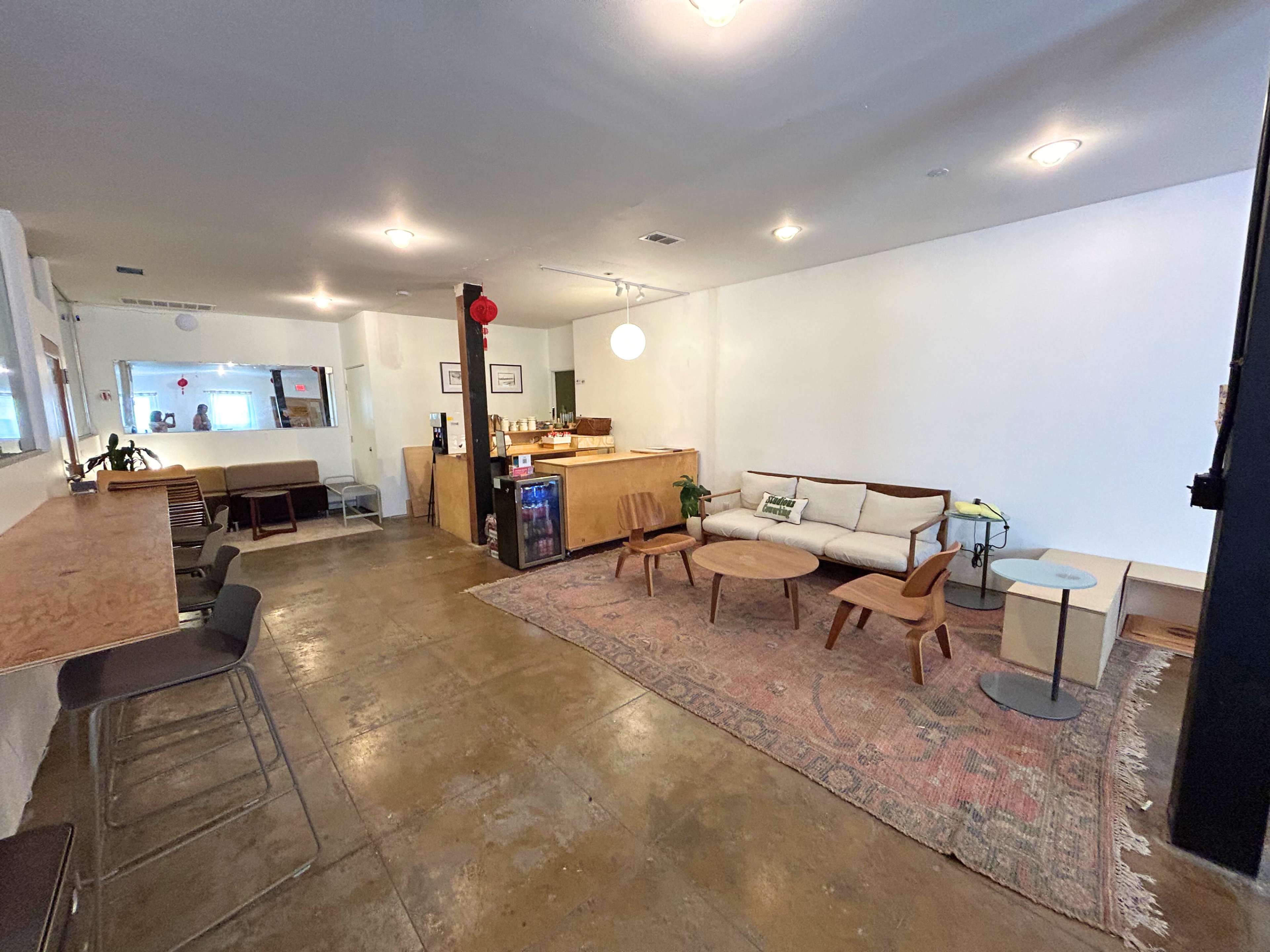 The image shows a modern, minimalist interior of a café with wooden furniture and a decorative rug on the concrete floor.