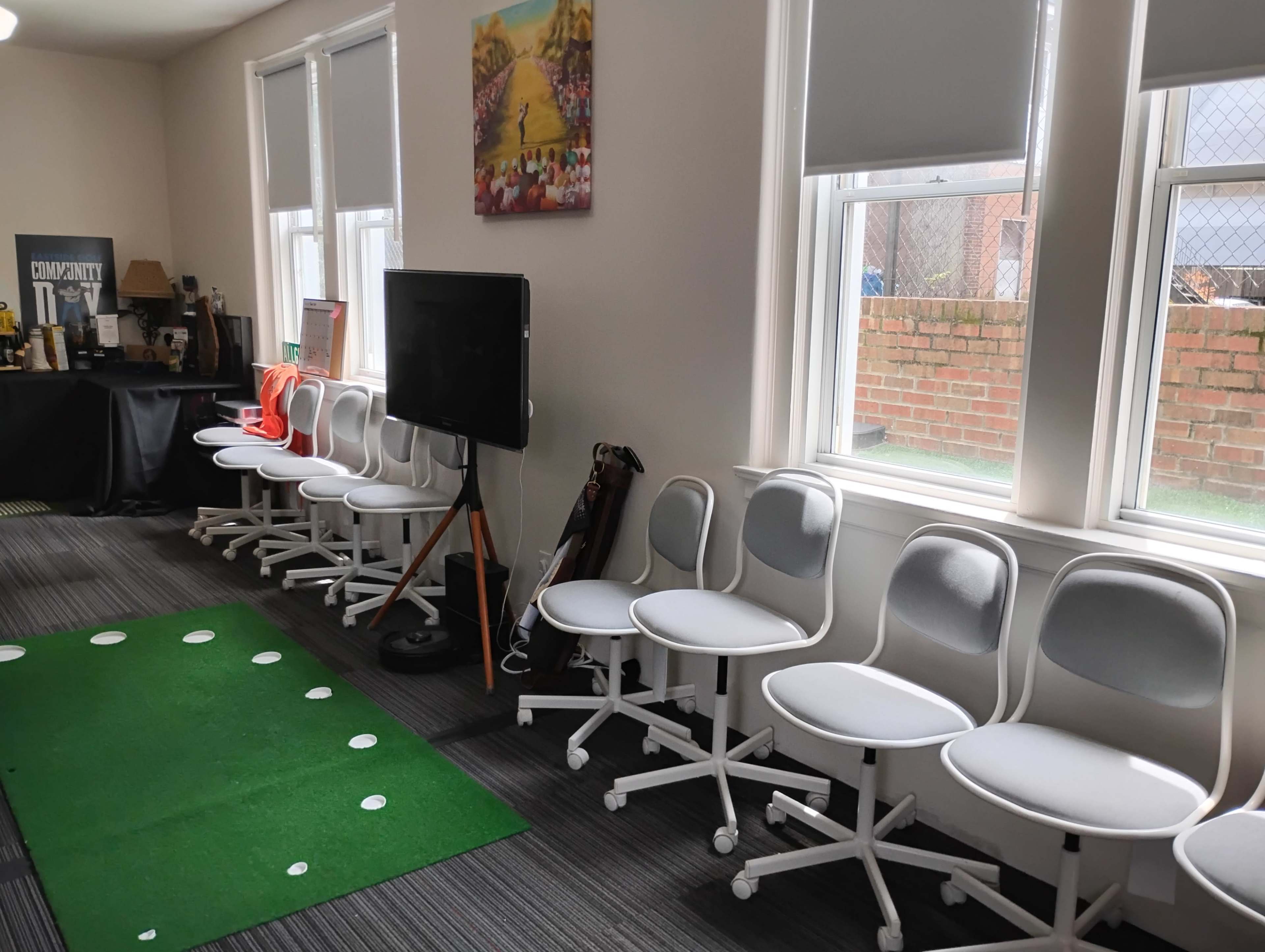 The image shows a room with a row of white office chairs, a television on a stand, and a green putting mat on the floor.
