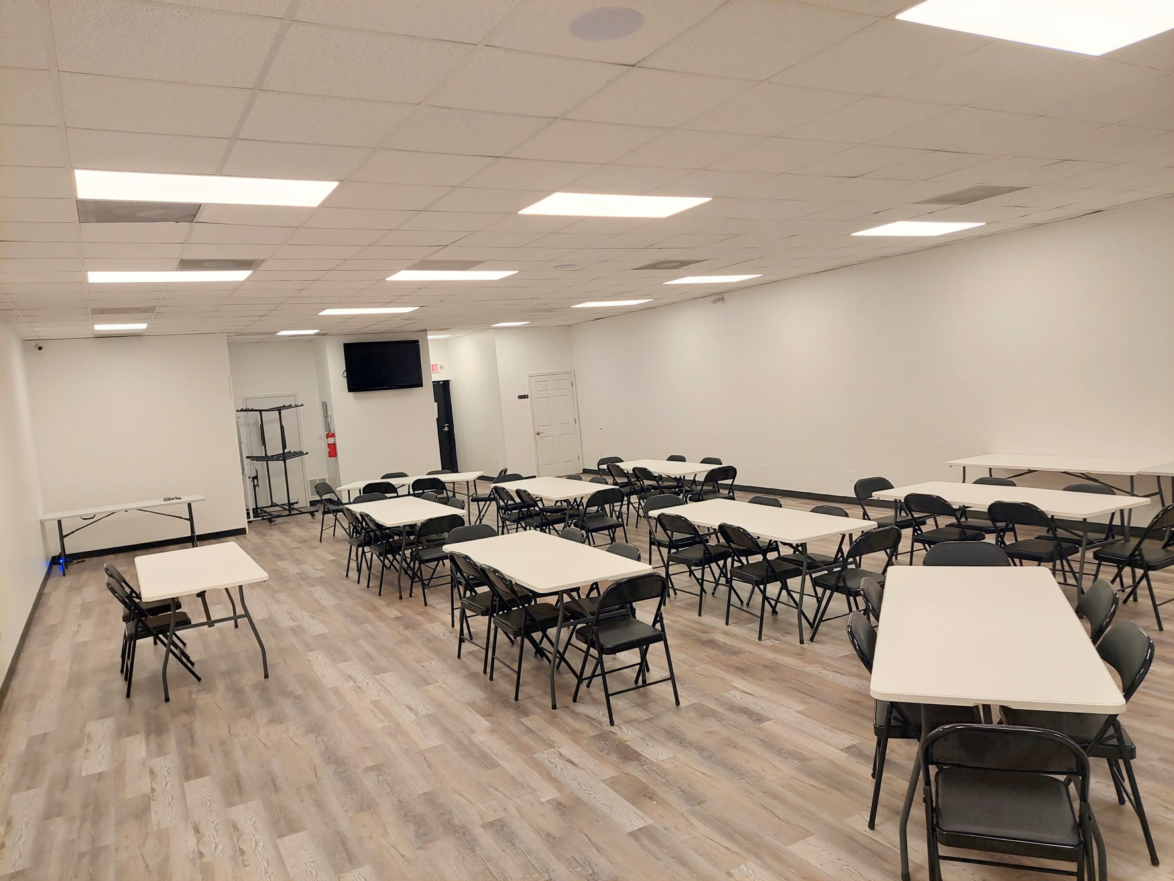 A spacious room with multiple tables and chairs arranged for a meeting or event, featuring a TV mounted on the wall.