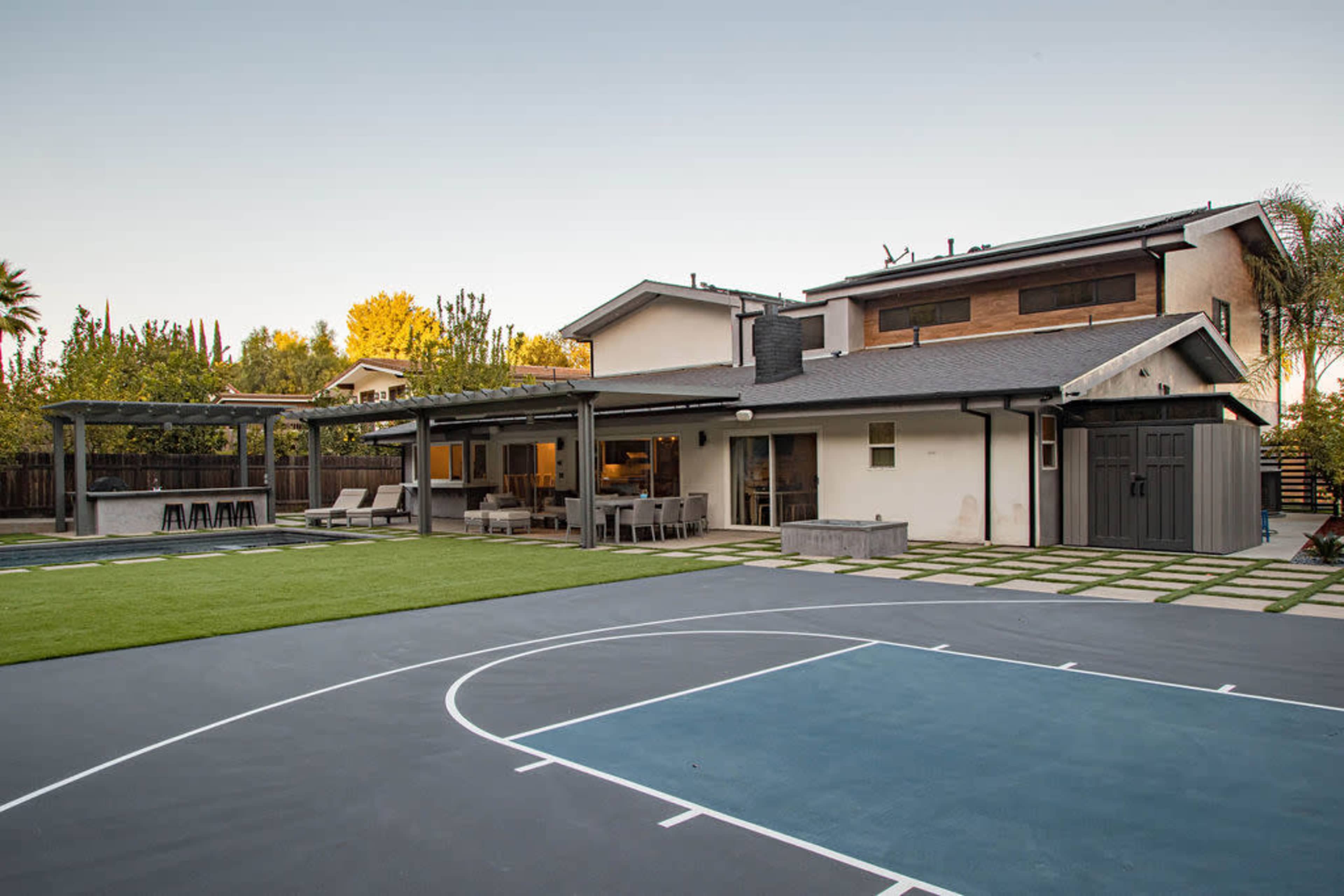 A modern backyard featuring a basketball court, a patio area with seating, and an exterior view of a two-story home.
