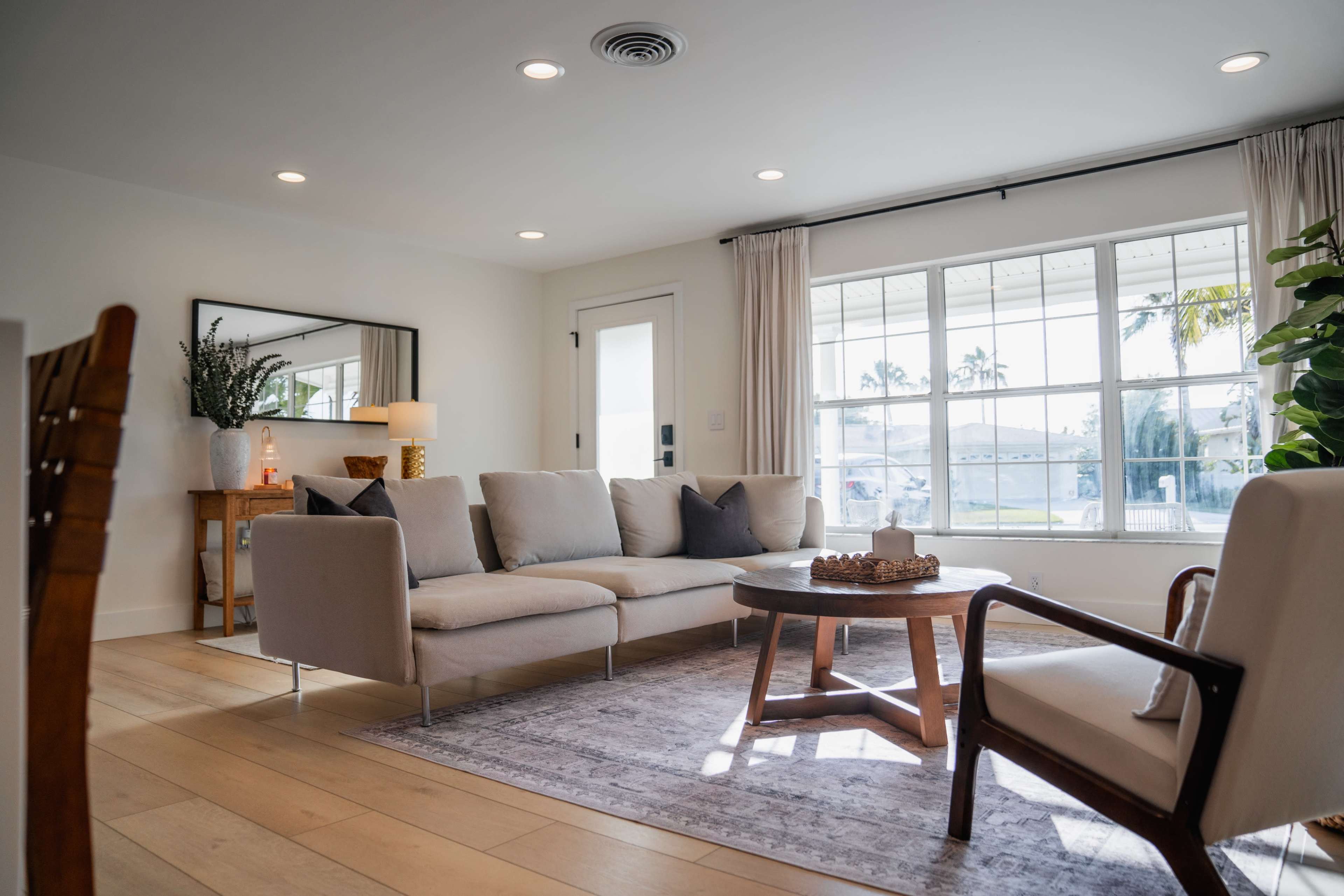The image shows a modern living room featuring a light-colored sectional sofa, a round coffee table, and large windows allowing natural light to enter.