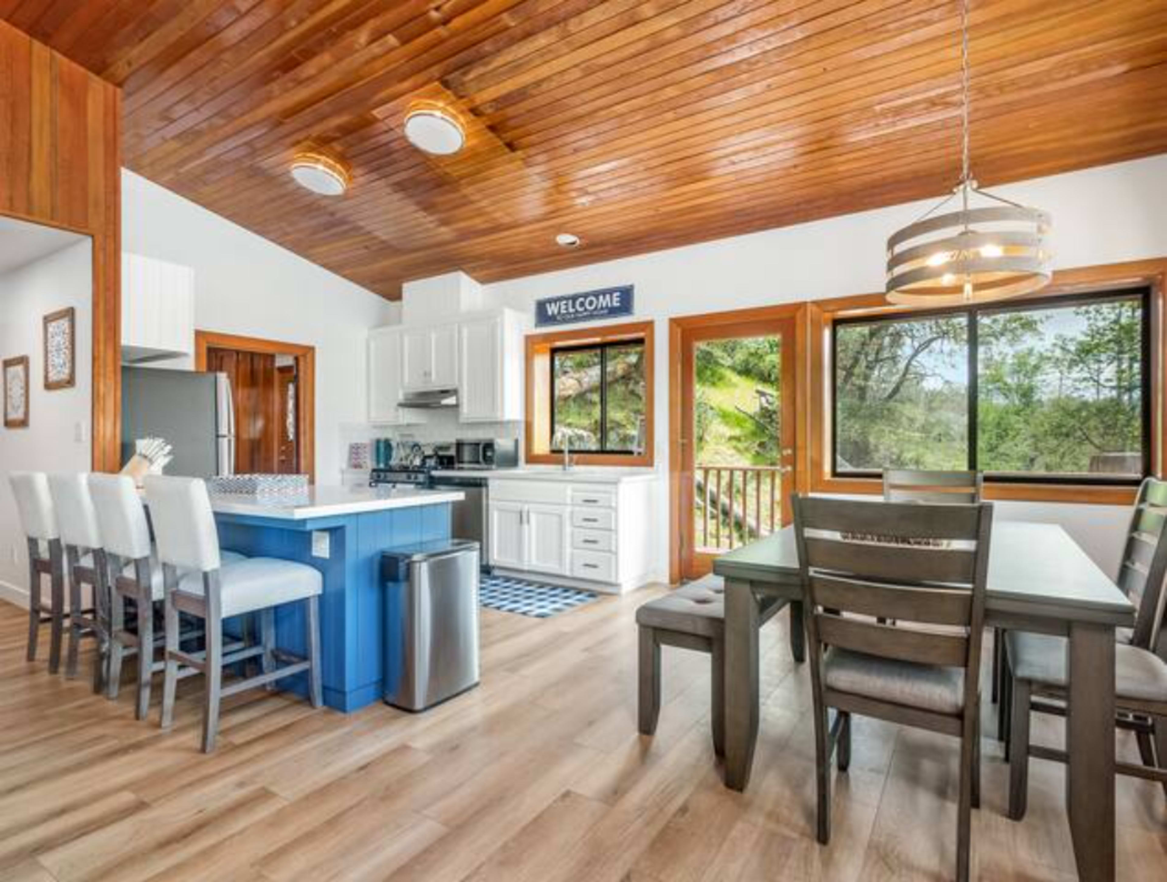 A modern kitchen and dining area features a wooden ceiling, white cabinetry, and a blue island surrounded by bar stools.
