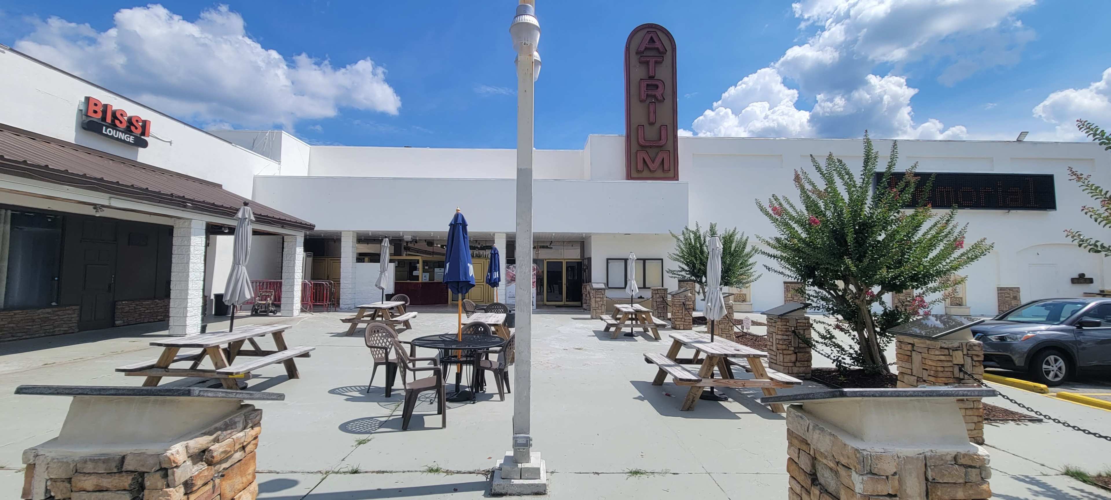 The image shows an outdoor seating area in front of a building labeled "ATRIUM," featuring wooden picnic tables and a blue umbrella, with a parking lot and decorative landscaping.
