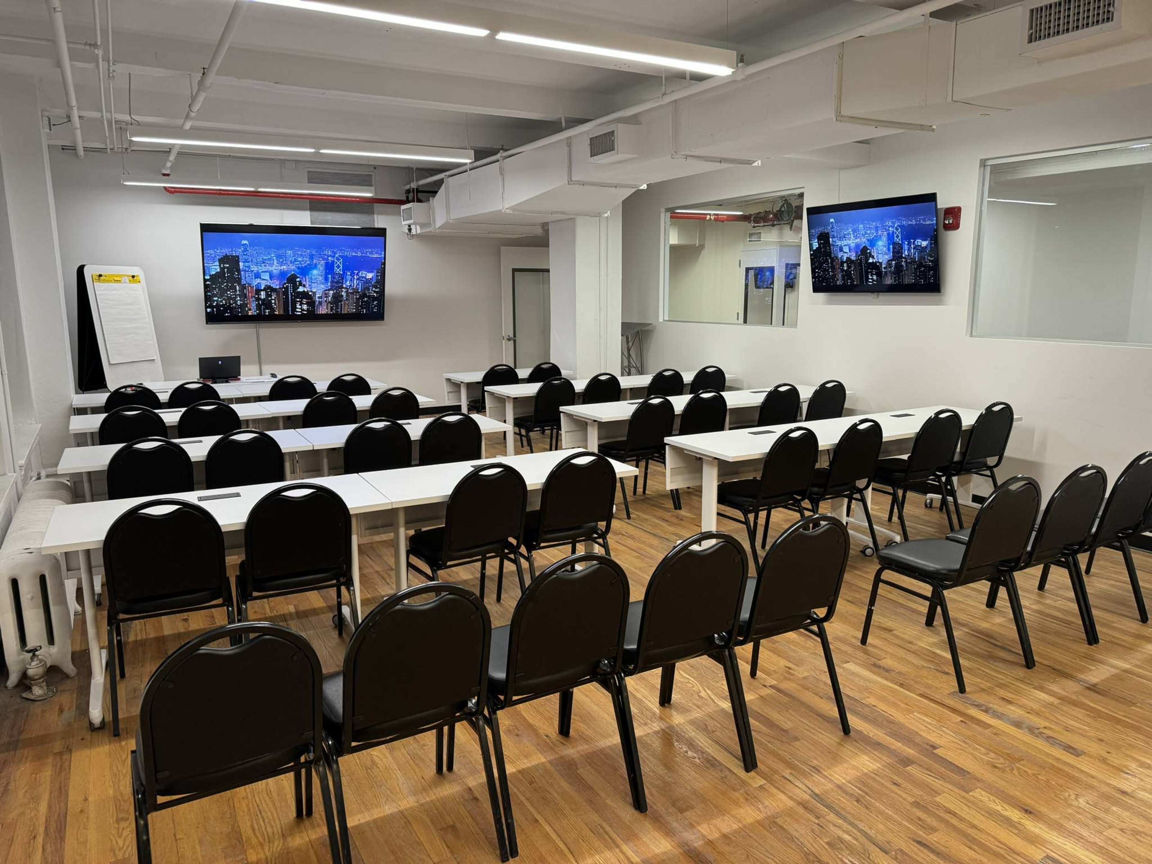 A meeting room features several rows of black chairs facing two wall-mounted screens displaying a city skyline.