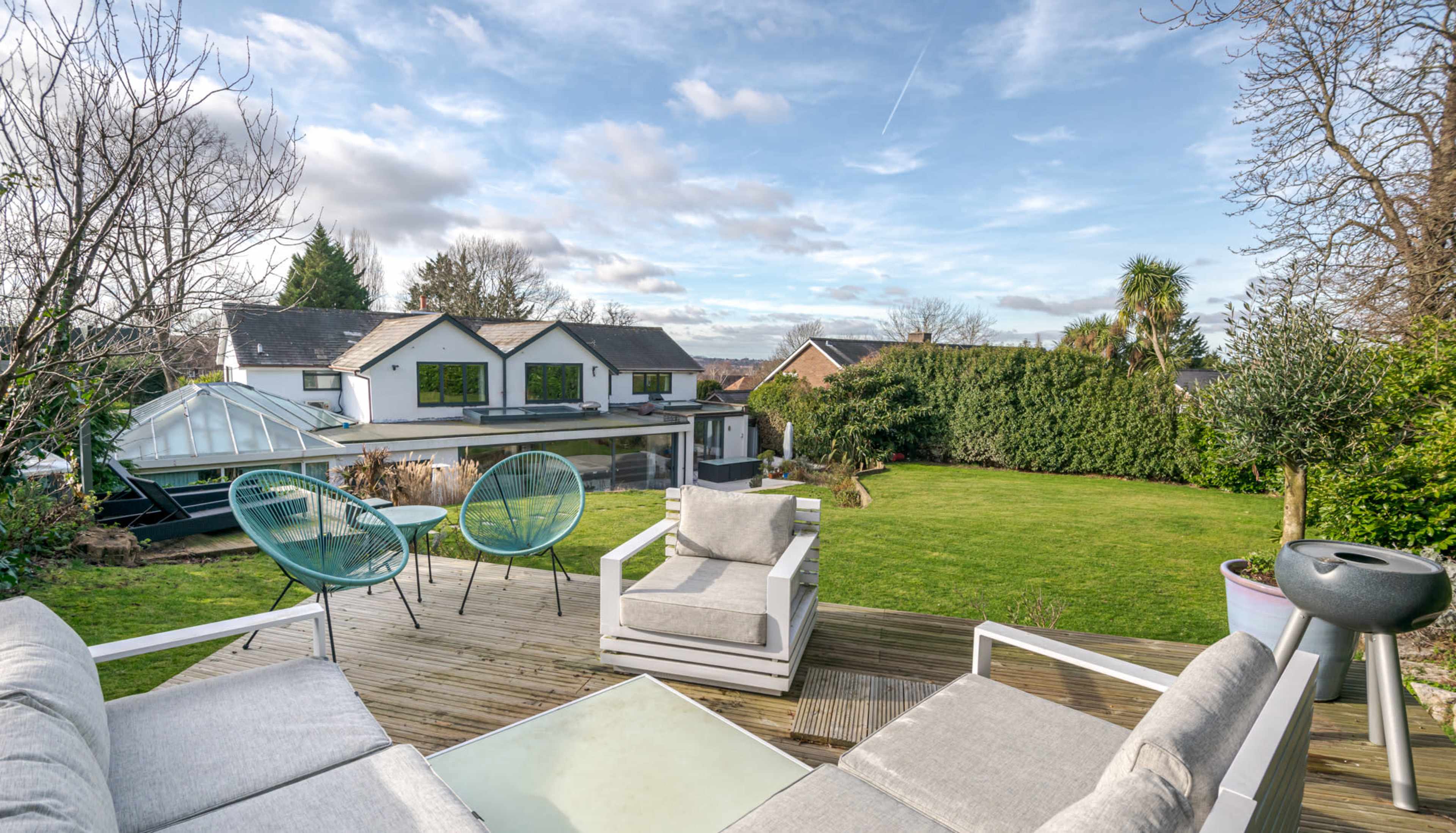 A wooden deck overlooks a landscaped garden and two houses, with modern outdoor furniture positioned for relaxation.