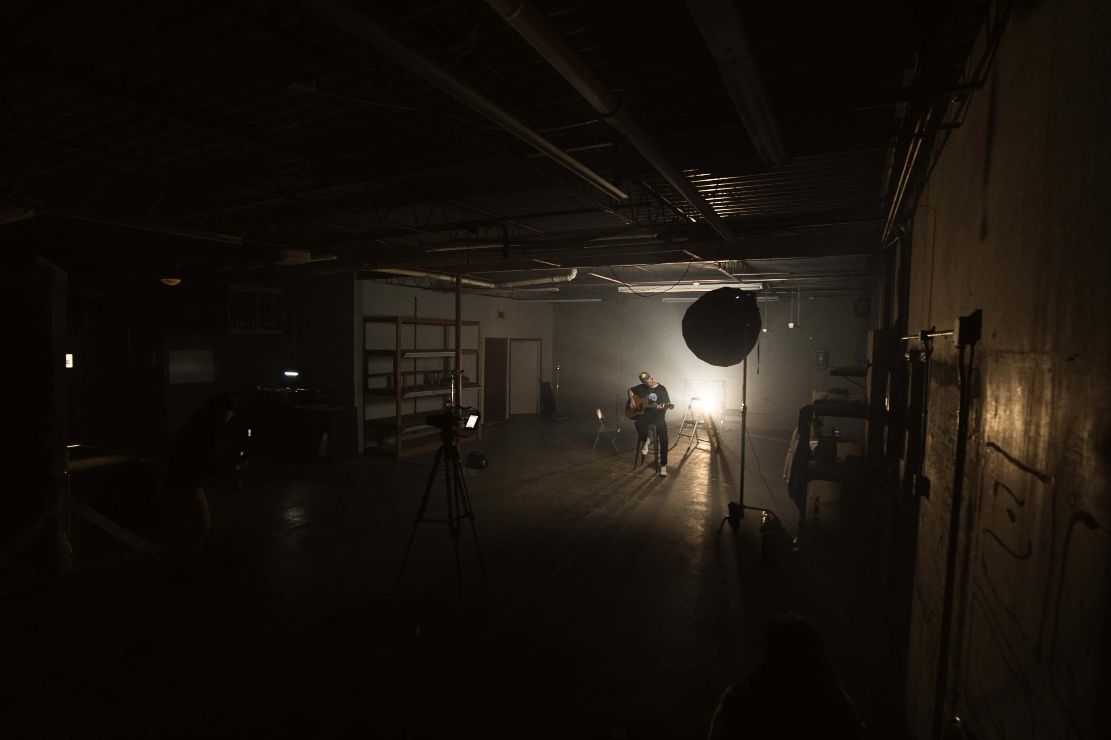 A person is sitting on a stool in a dimly lit warehouse, illuminated by a spotlight, while two cameras are positioned nearby.