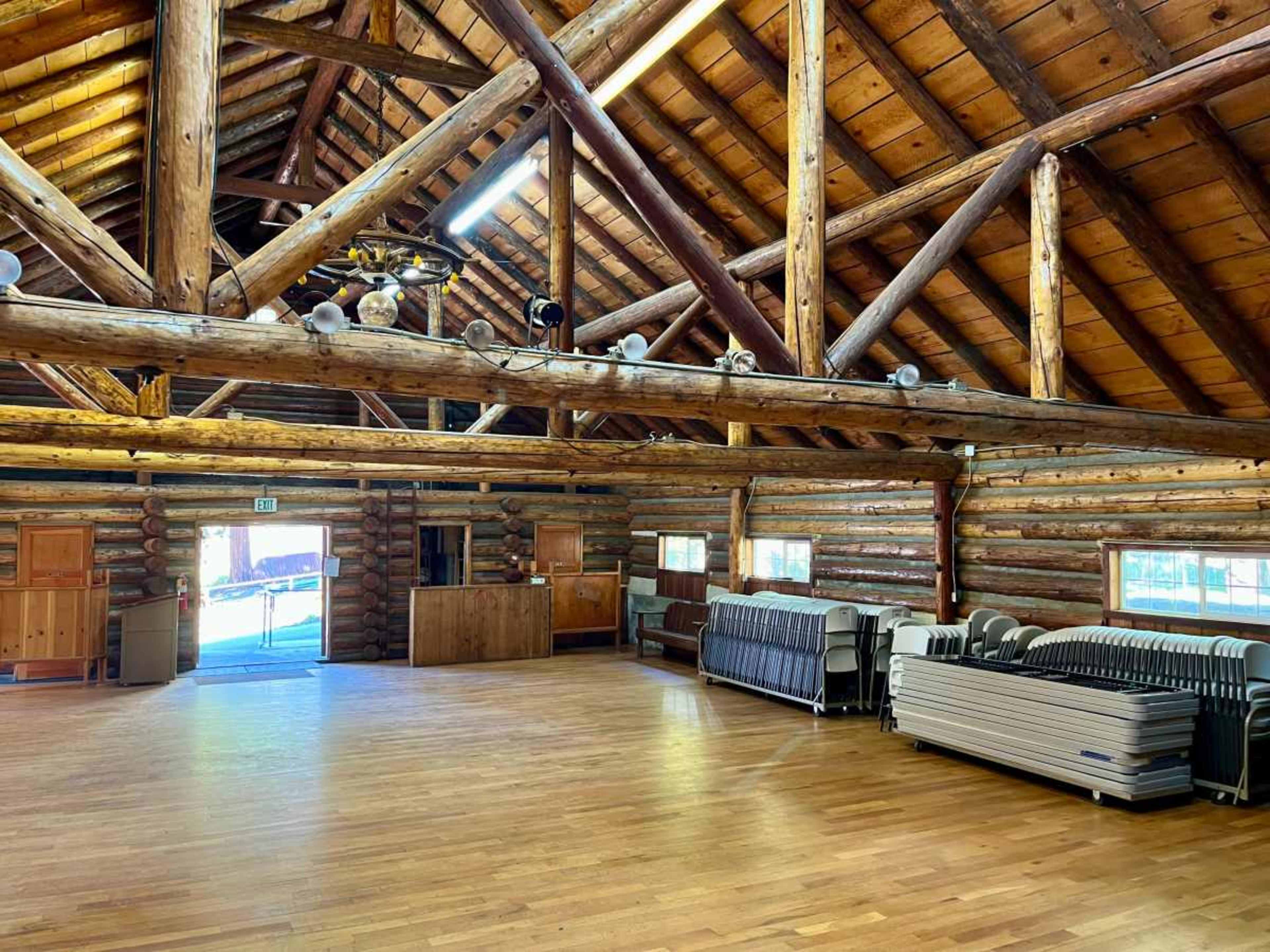 The interior of a spacious log cabin features exposed wooden beams and stacked logs, with folding tables stacked against the wall and large windows allowing natural light to enter.