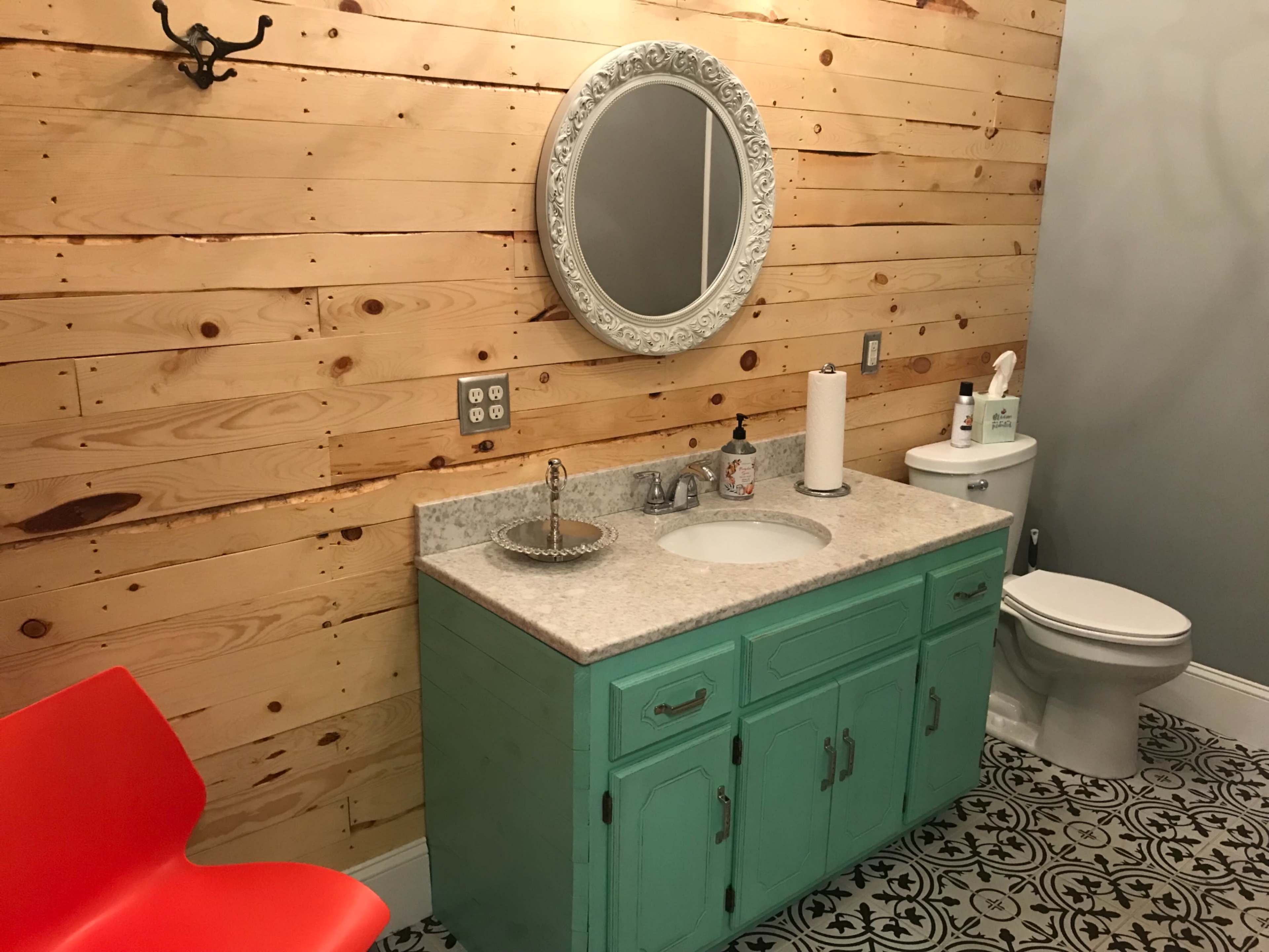 The image shows a bathroom featuring a light wood-paneled wall, a turquoise vanity with a countertop sink, a round mirror, a red chair, and a patterned tile floor.