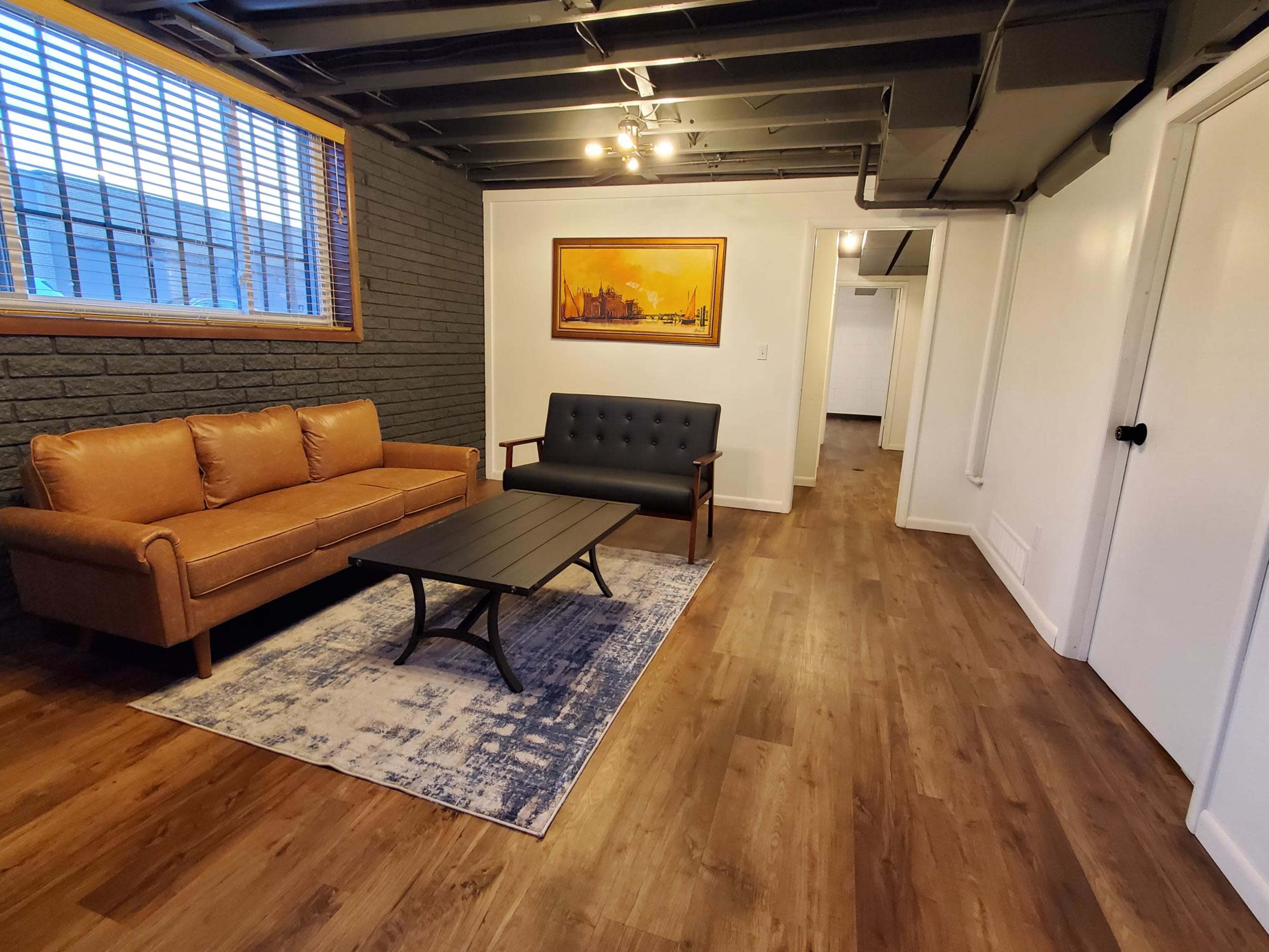 The image shows a modern, finished basement living area featuring a brown leather sofa, a black upholstered chair, a coffee table, and a decorative area rug.