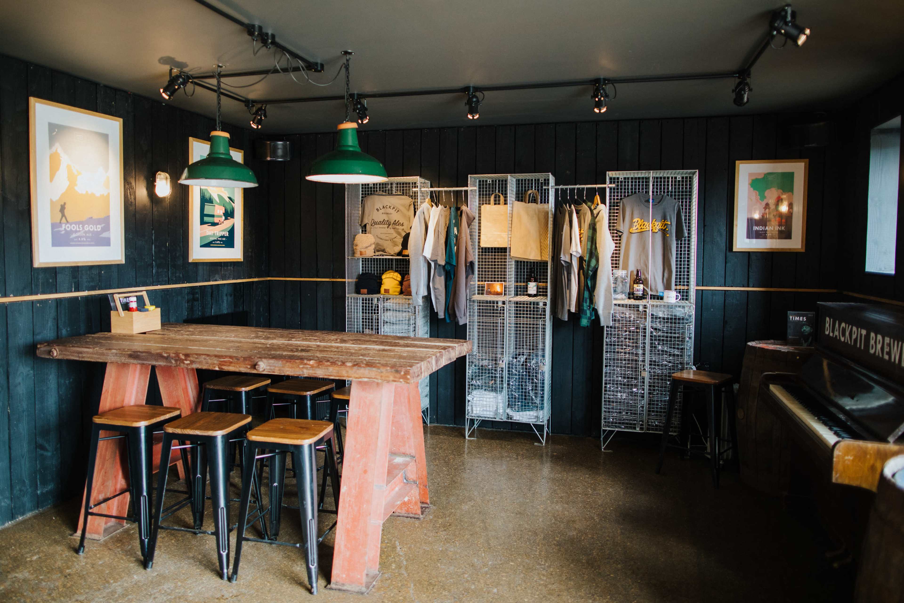 A rustic interior of a shop with a large wooden table, metal stools, and clothing displayed on wire shelving.