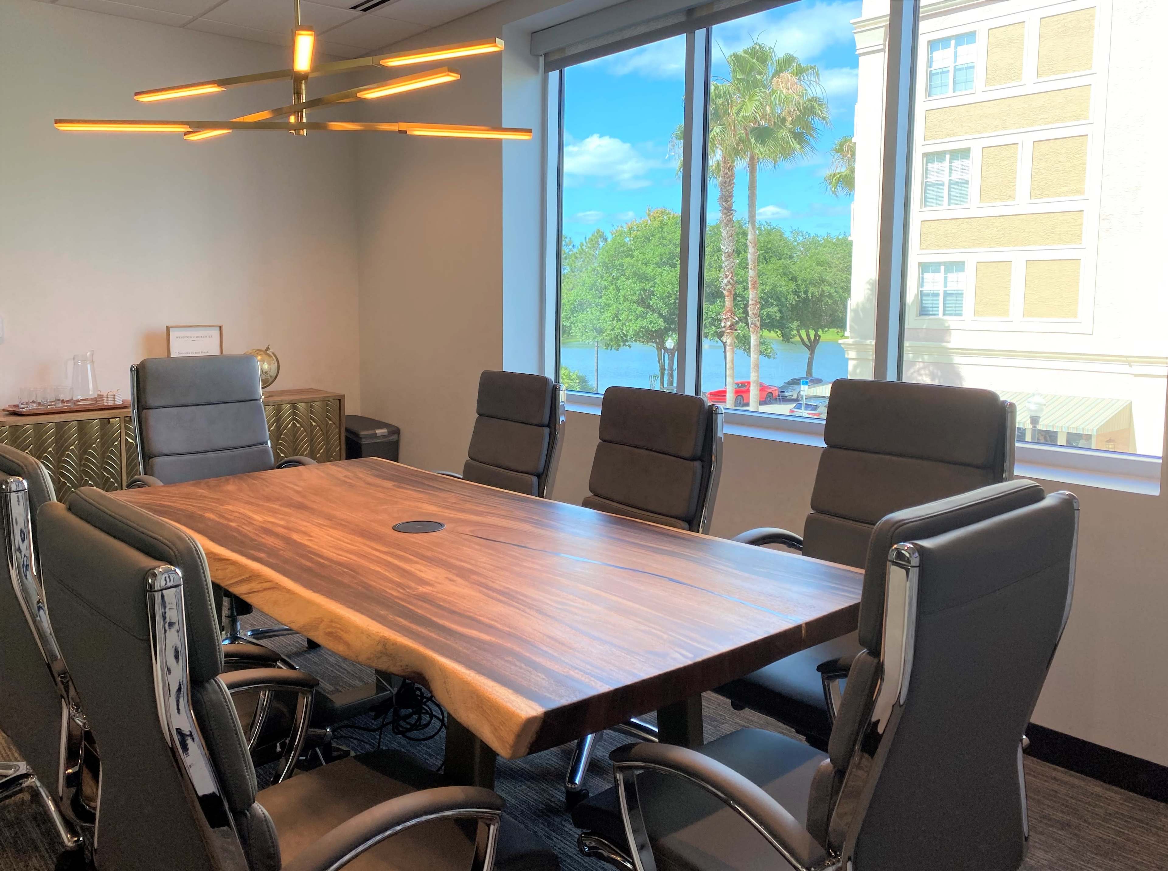 A large wooden conference table with ergonomic chairs is set up in a well-lit meeting room, featuring a view of palm trees and a building through a large window.