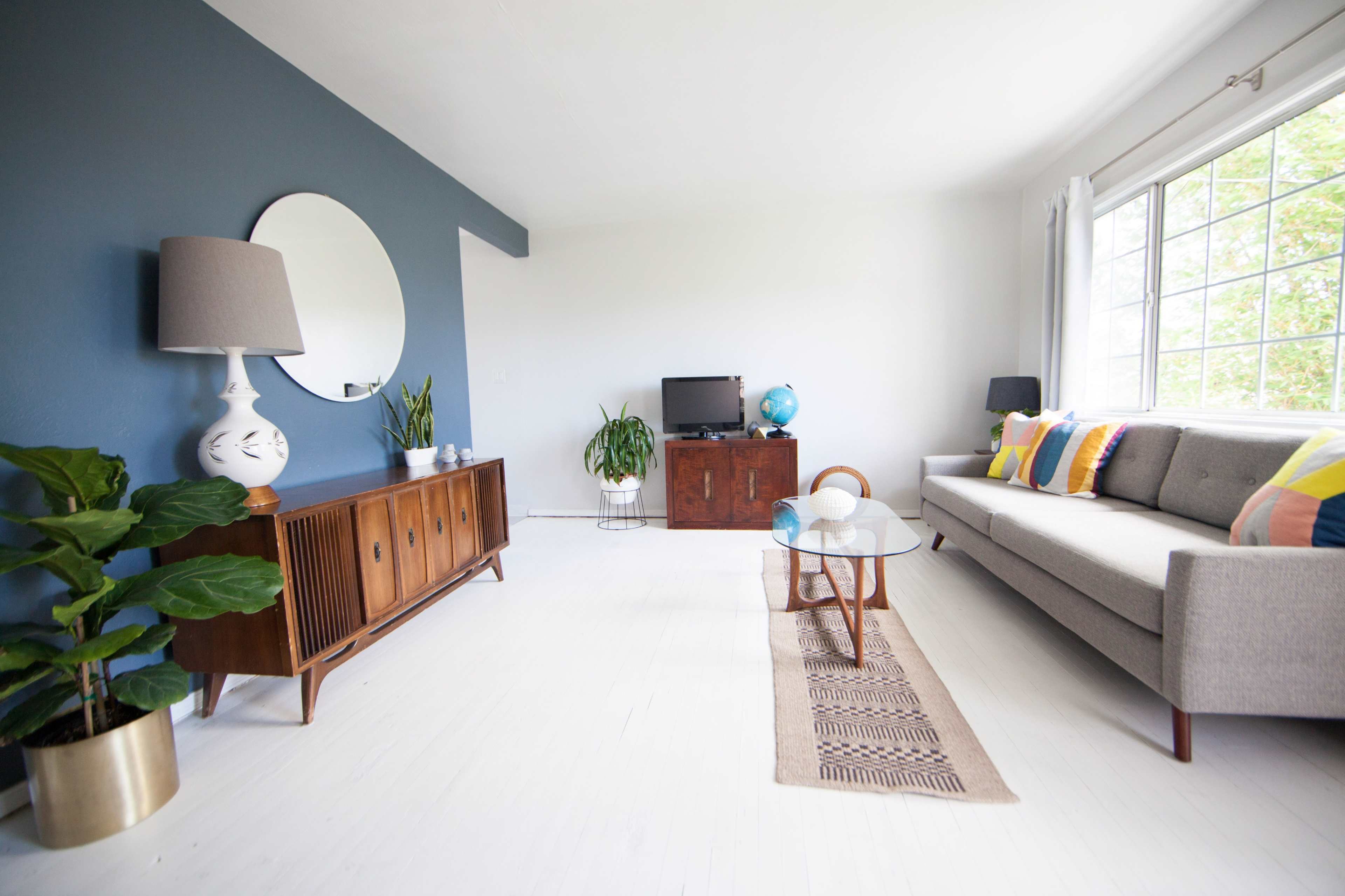 The living room features a gray sofa, a wooden coffee table, and a media console against a two-tone wall, with a large window letting in natural light.