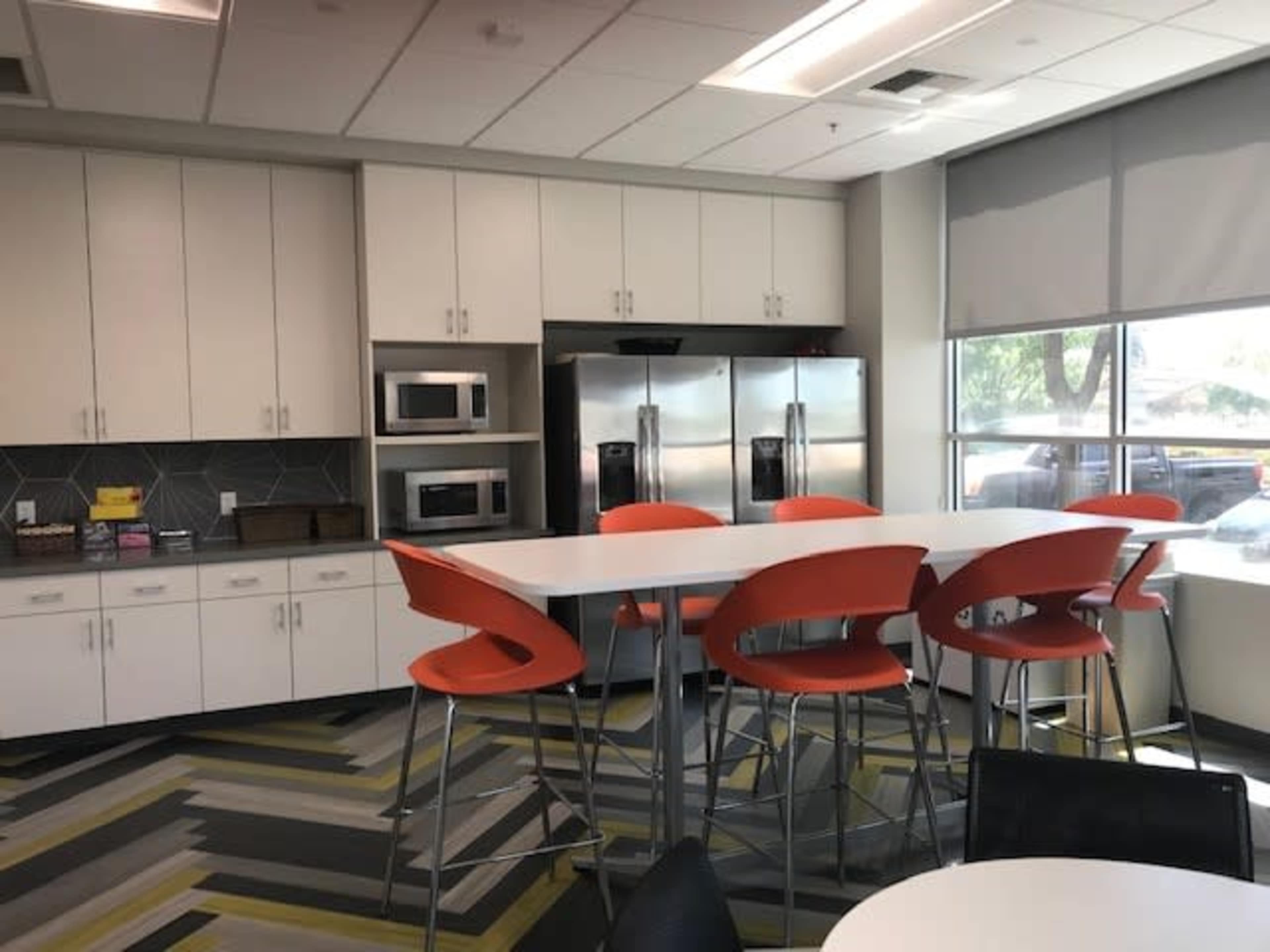 A modern kitchen area with white cabinets, stainless steel appliances, and orange bar stools around a long table.
