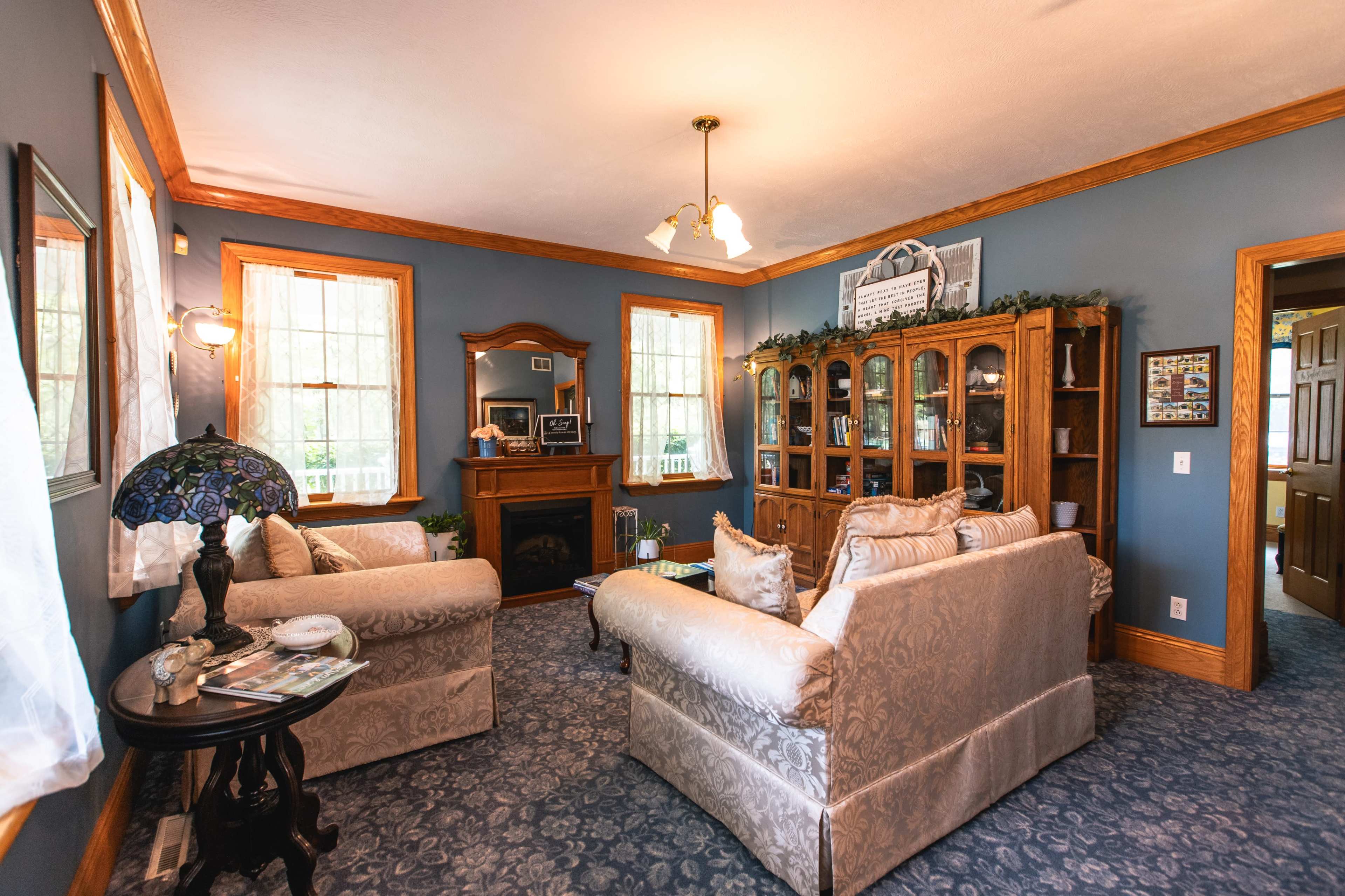 The living room features two sofas facing a wooden cabinet, with a fireplace and framed mirrors against blue walls adorned with windows.
