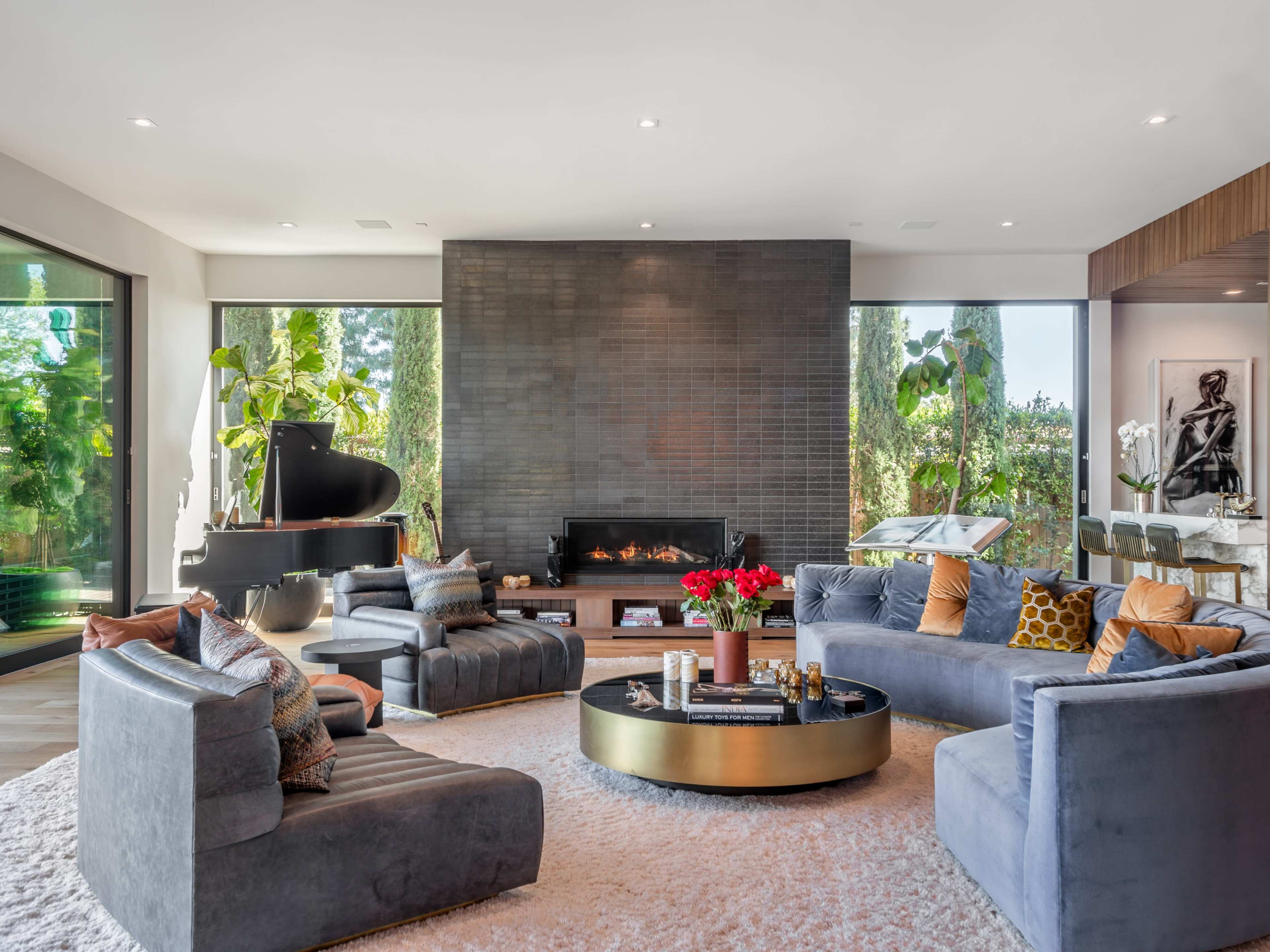 A contemporary living room features a large, circular coffee table surrounded by grey sectional sofas, with a fireplace and large windows showcasing greenery outside.