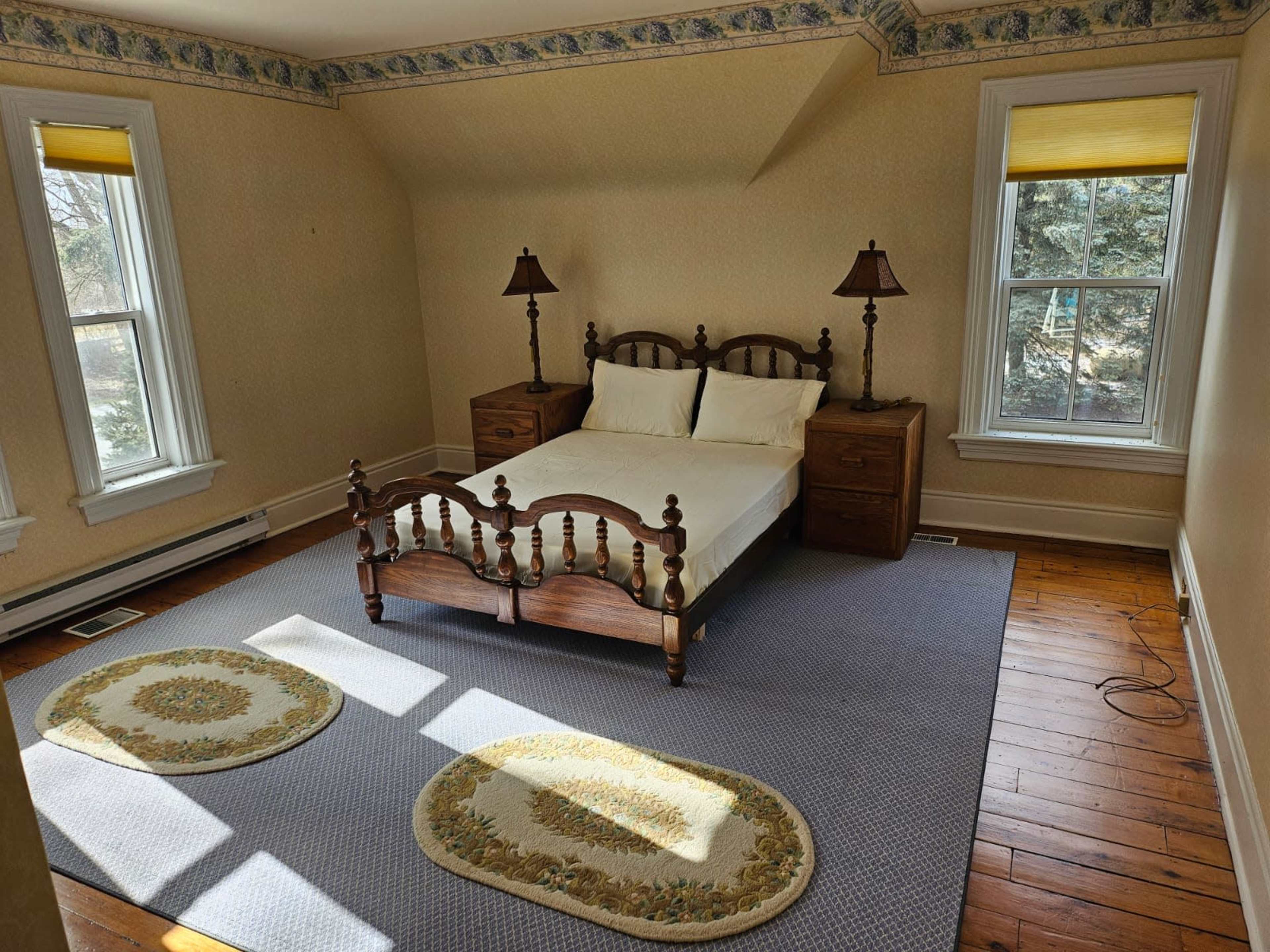 A sparsely furnished bedroom with a wooden bed frame, two lamps on nightstands, and two windows allowing natural light to enter.