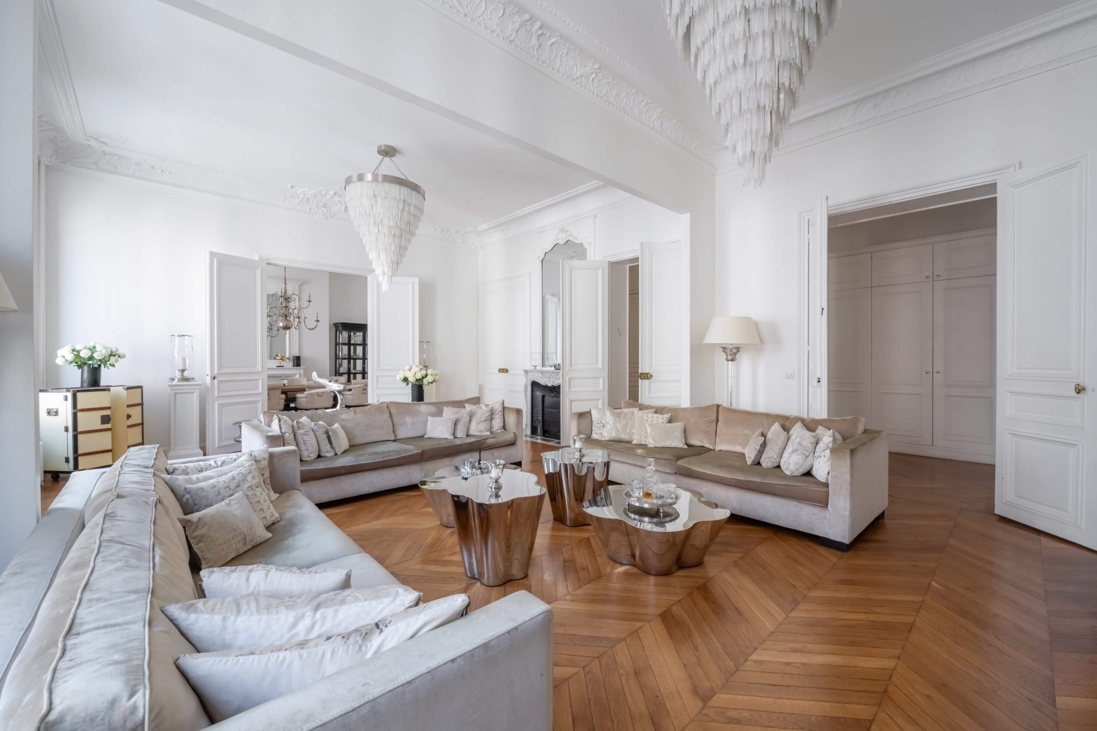 A spacious living room features two light gray sofas with decorative pillows, a pair of modern side tables, and a chandelier, all set against a backdrop of elegant white walls and hardwood flooring.