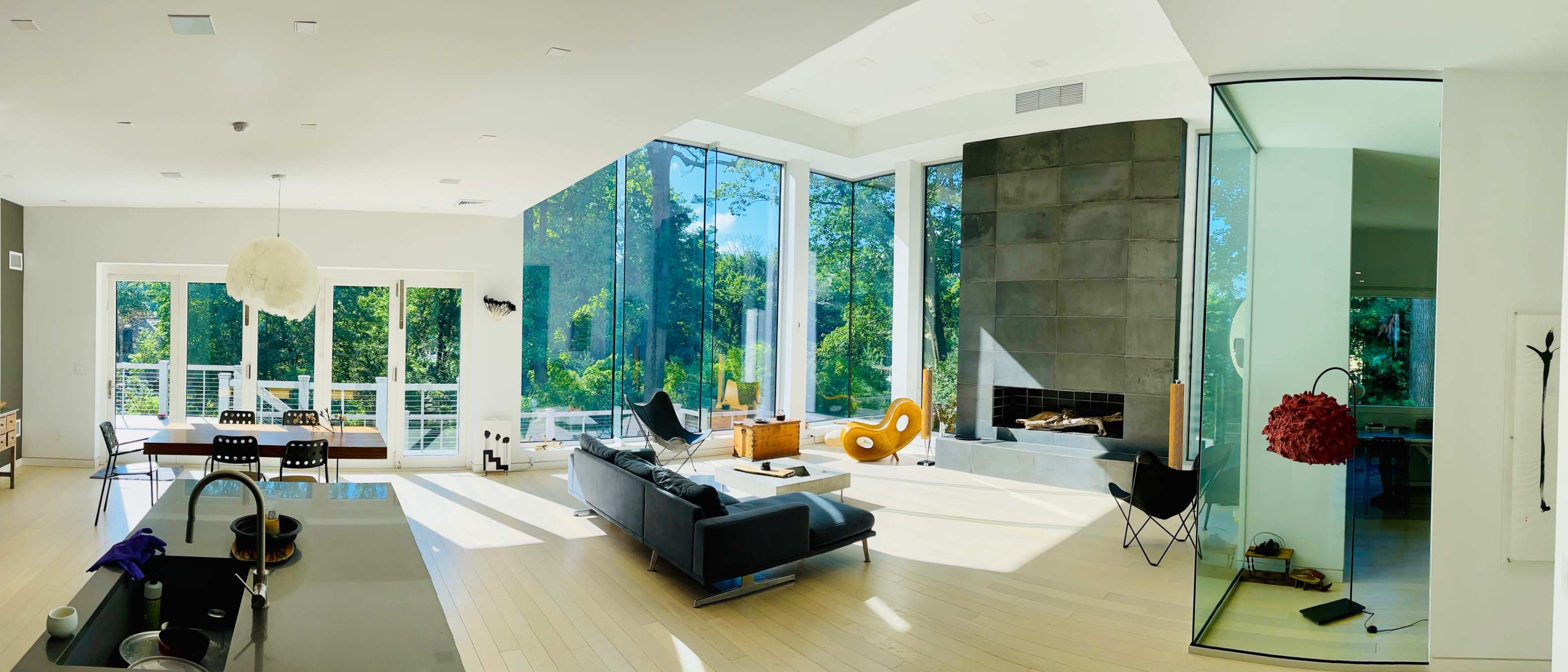 A spacious living room with large glass windows overlooking greenery, featuring a sofa, a modern chair, a fireplace, and a dining area.