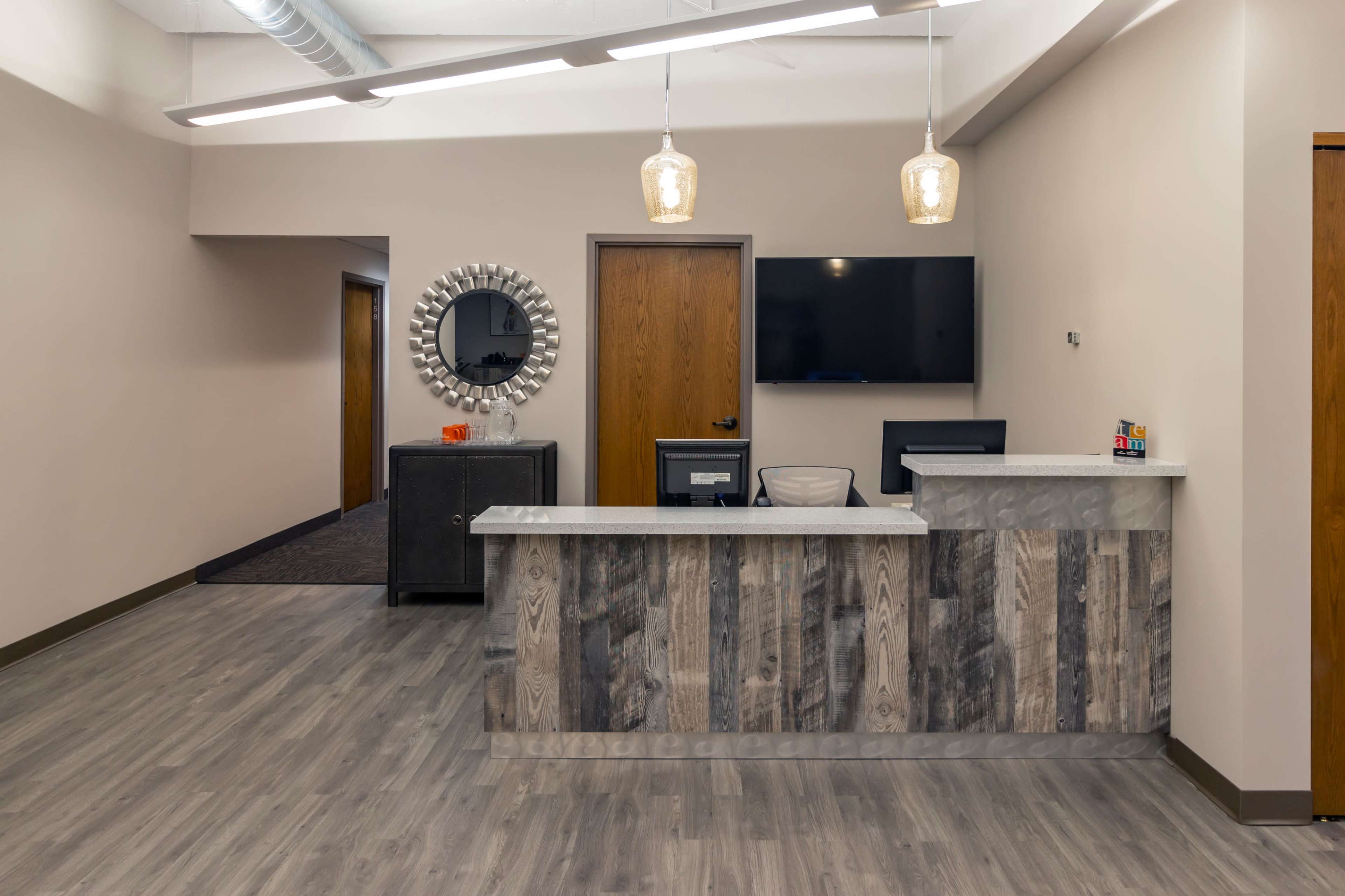 A modern reception area featuring a wood and marble front desk, pendant lighting, a large circular mirror, and a wall-mounted television.