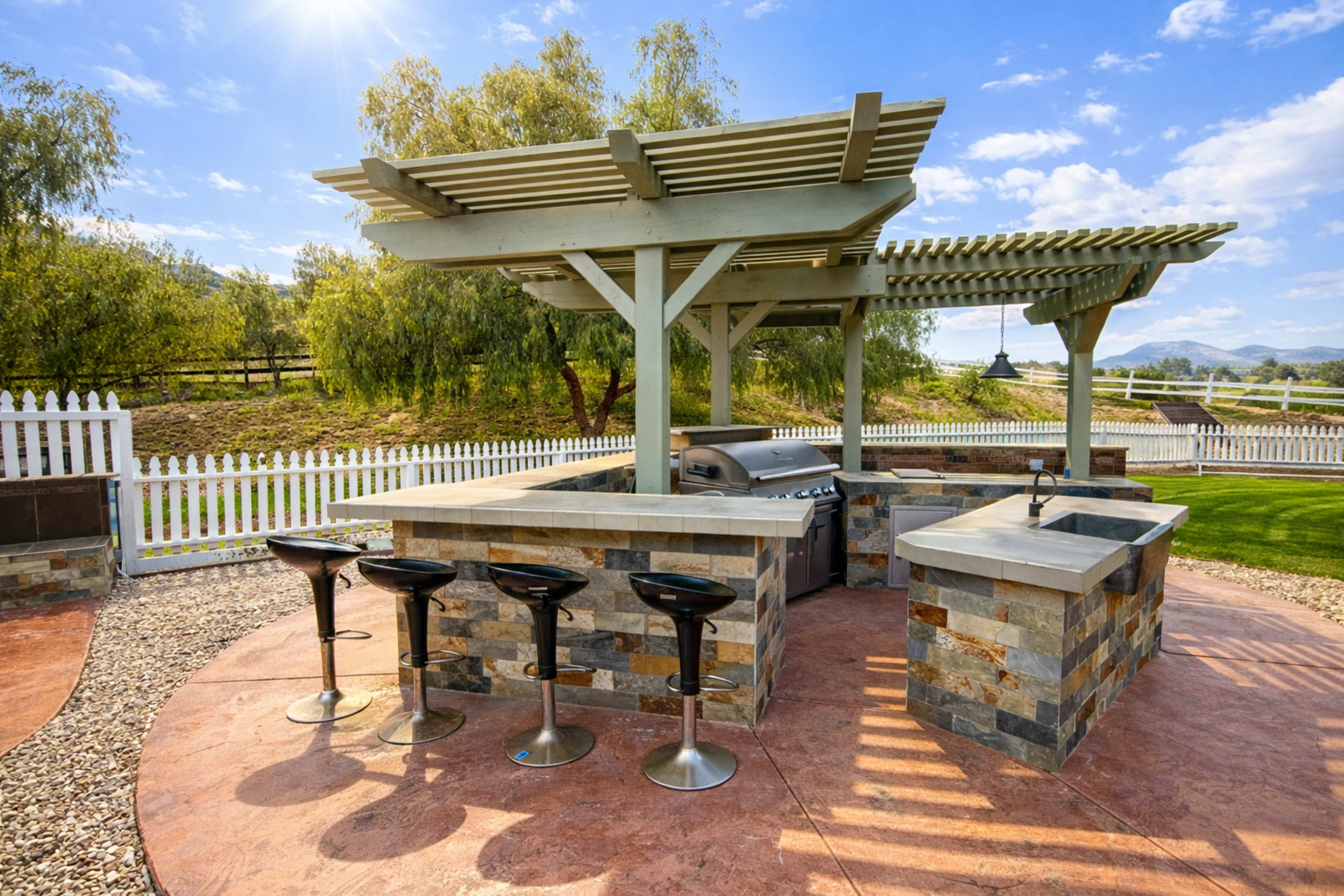 The image shows an outdoor kitchen with a grill, bar seating, and a stone countertop situated in a landscaped yard.