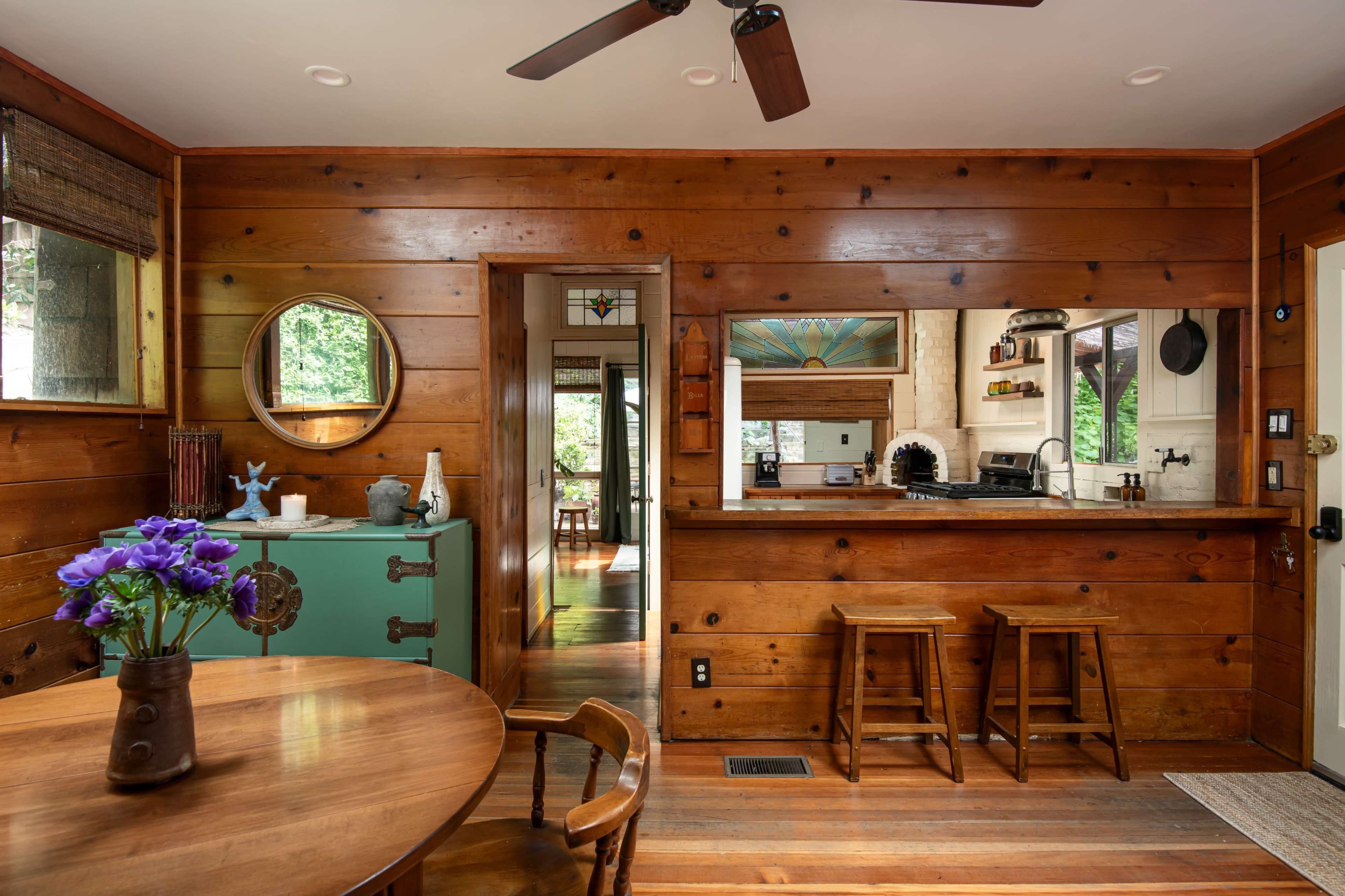 The image shows a rustic wooden interior featuring a round table with chairs, a green cabinet, and a kitchen visible through an open space with stools.