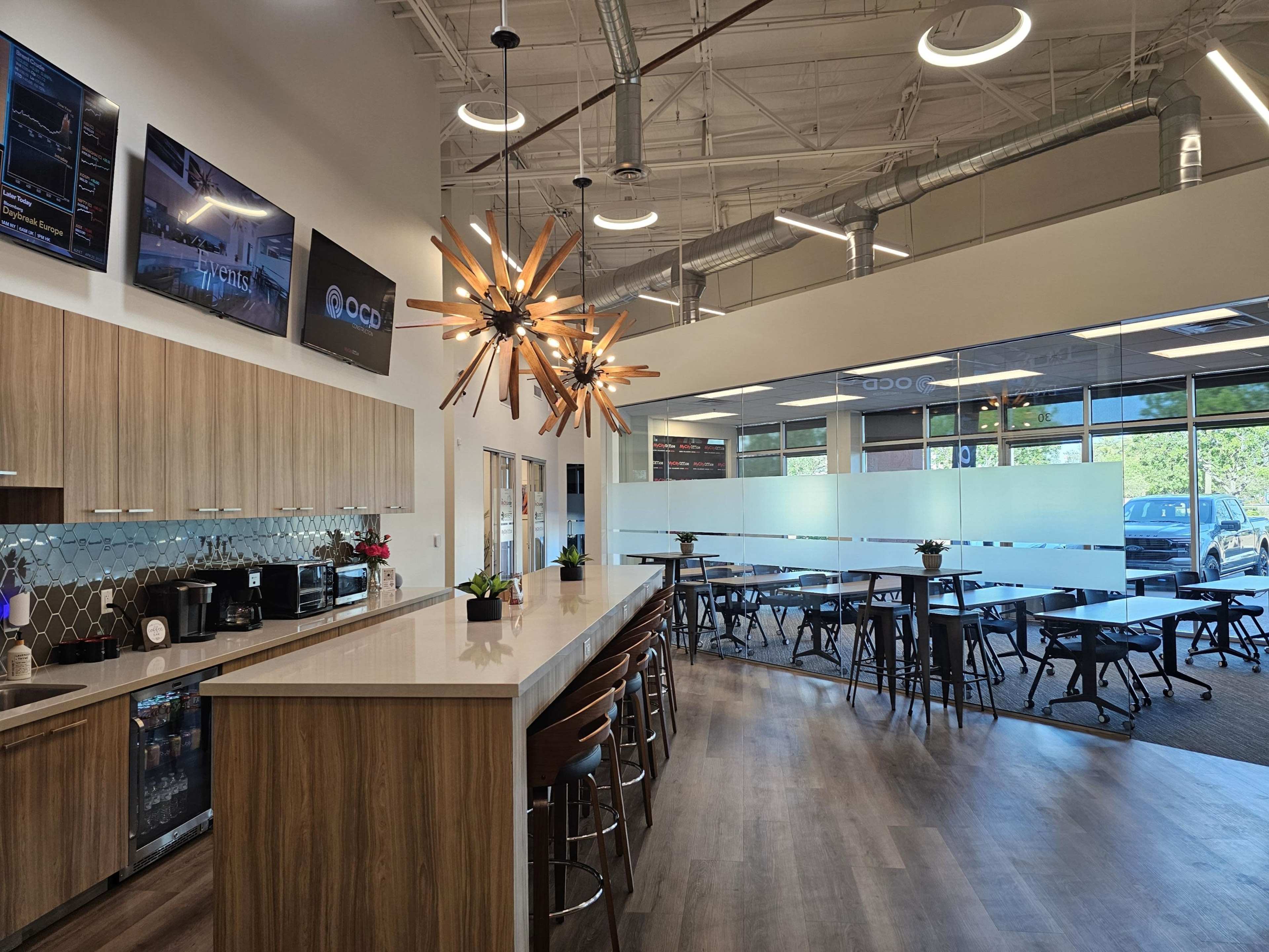 The image shows a modern office kitchen area with a long wooden counter, a dining space featuring several tables, and large windows allowing natural light.