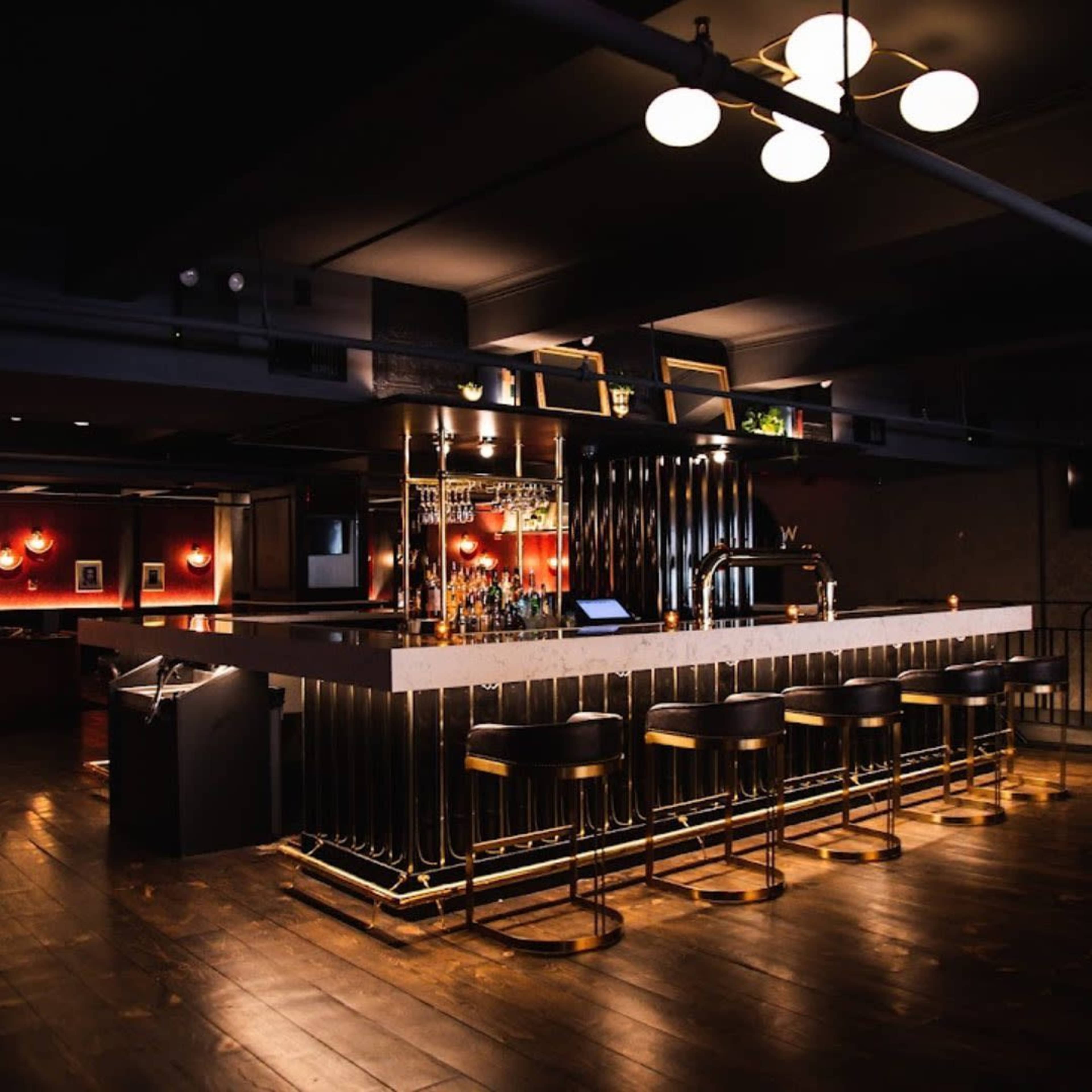 The image shows a modern bar with a sleek design featuring a marble countertop, black stools, and warm lighting in a dimly lit setting.