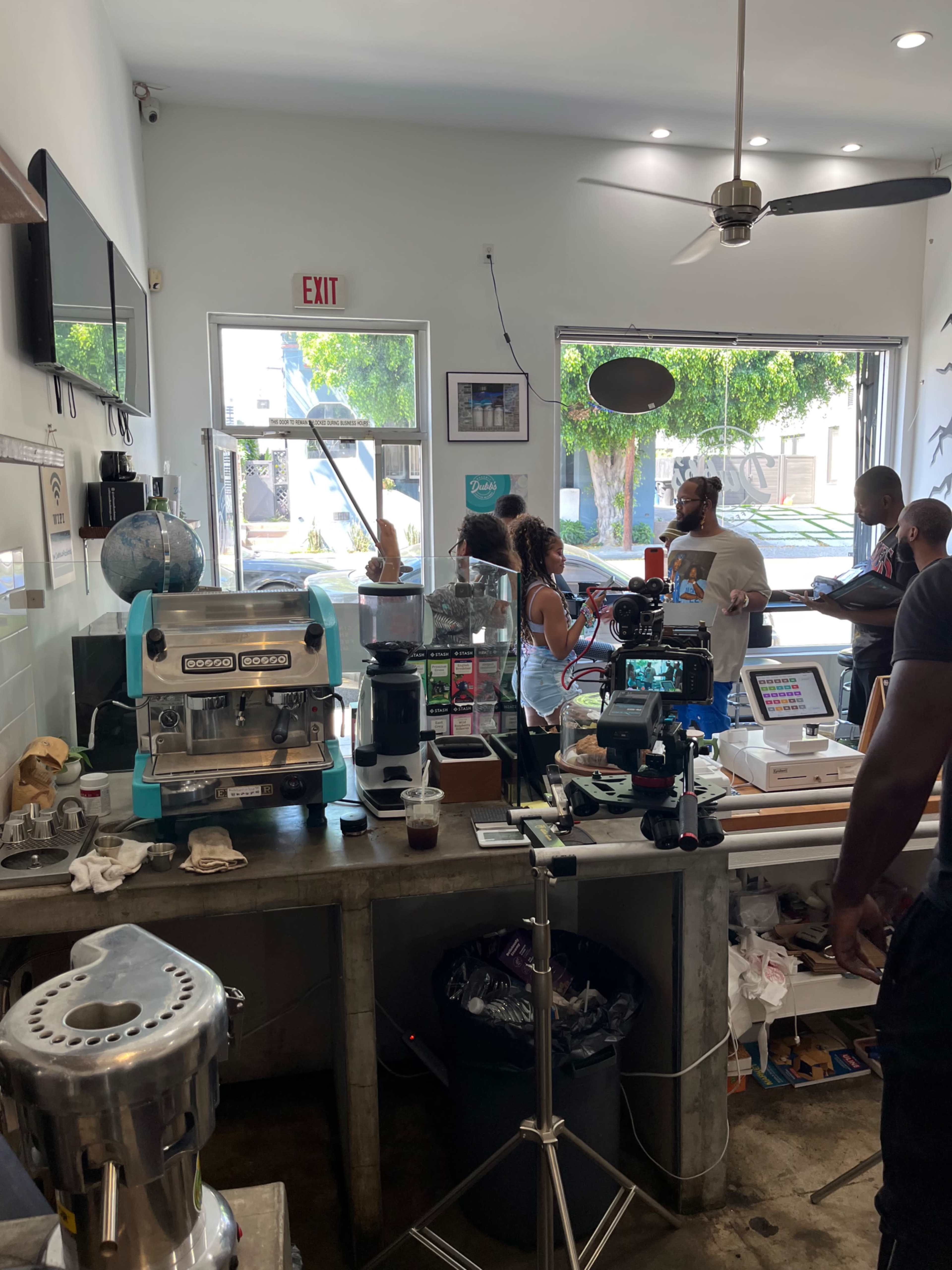 The image shows a coffee shop with a barista preparing drinks behind a counter while customers engage nearby, and filming equipment is set up in the foreground.