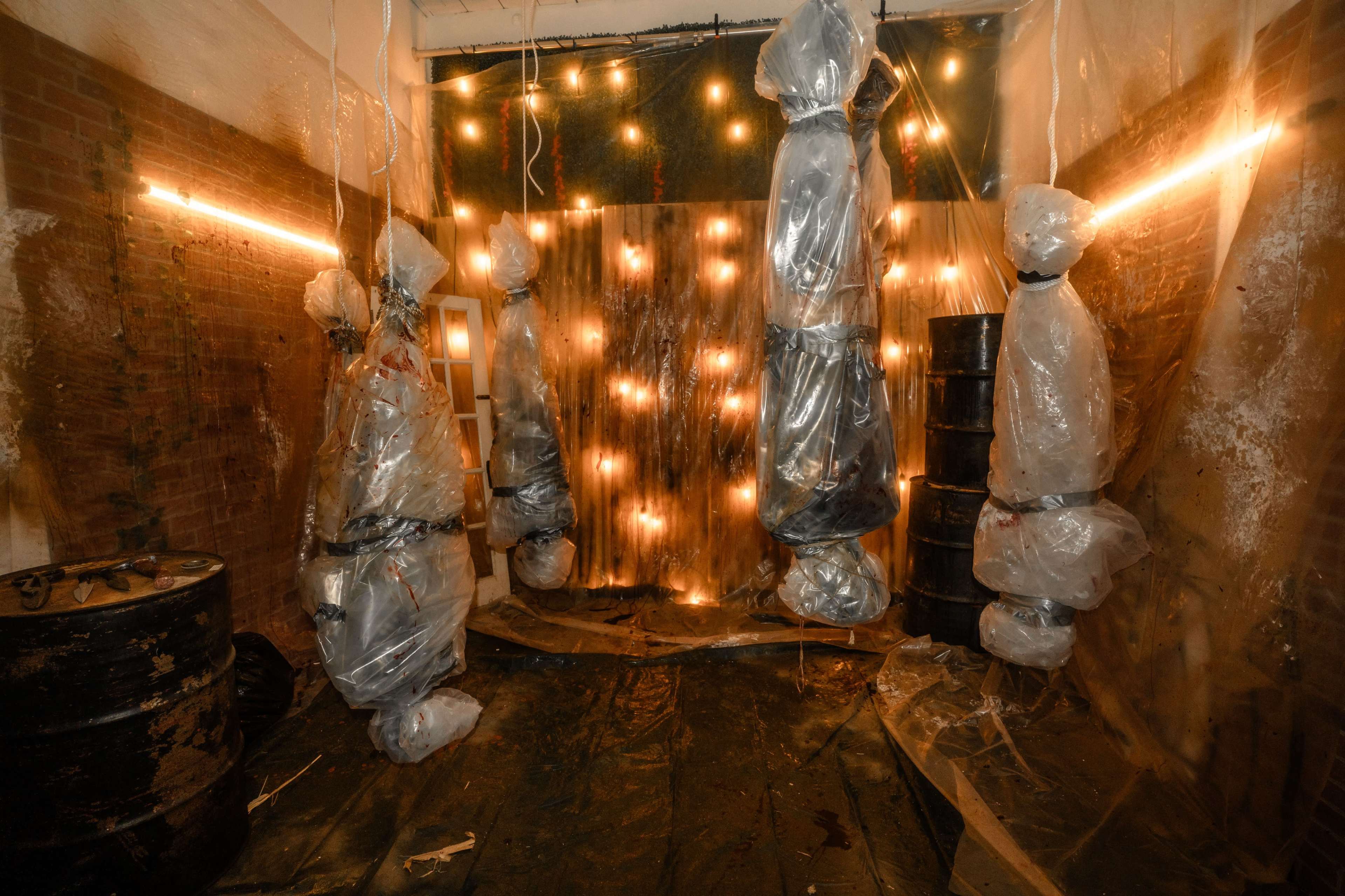 The image shows a dimly lit room with figures covered in plastic suspended from the ceiling, surrounded by barrels and string lights on the walls.