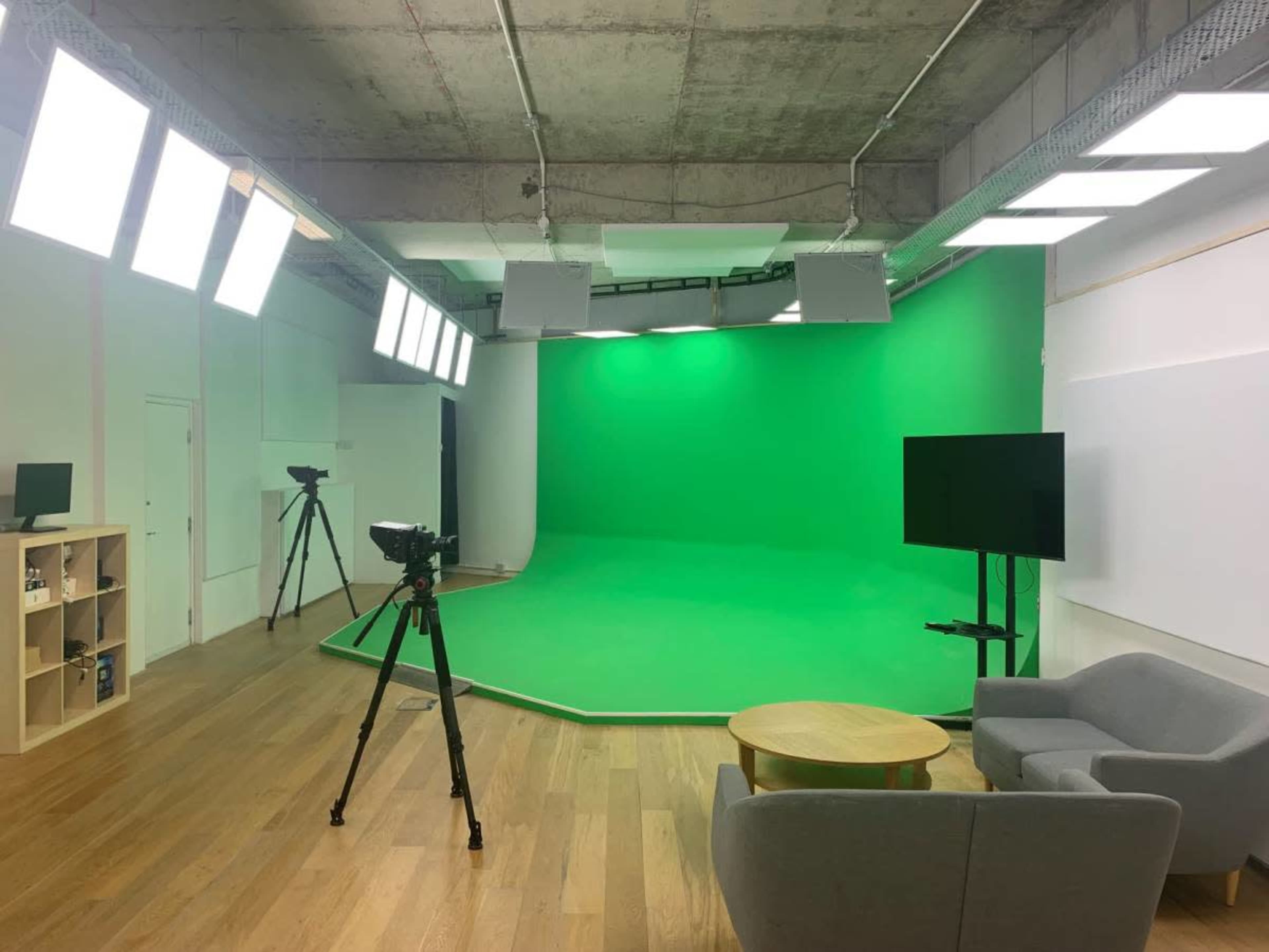 The image shows a green screen studio equipped with cameras, lighting fixtures, a television, and seating arranged around a wooden table.