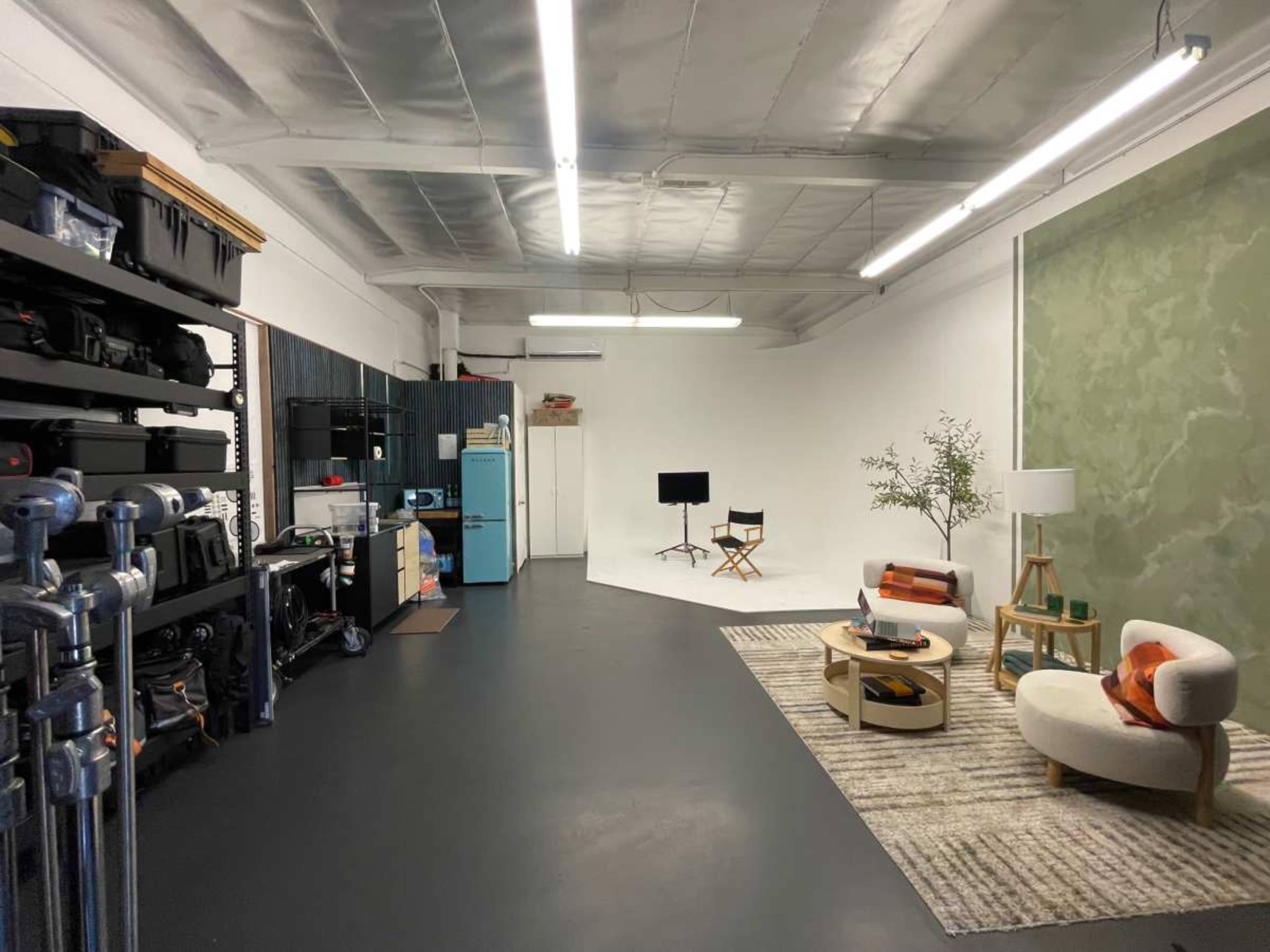 The image depicts a spacious studio with a minimalist design, featuring a seating area with a sofa, coffee table, and a lamp, alongside storage shelves filled with equipment.