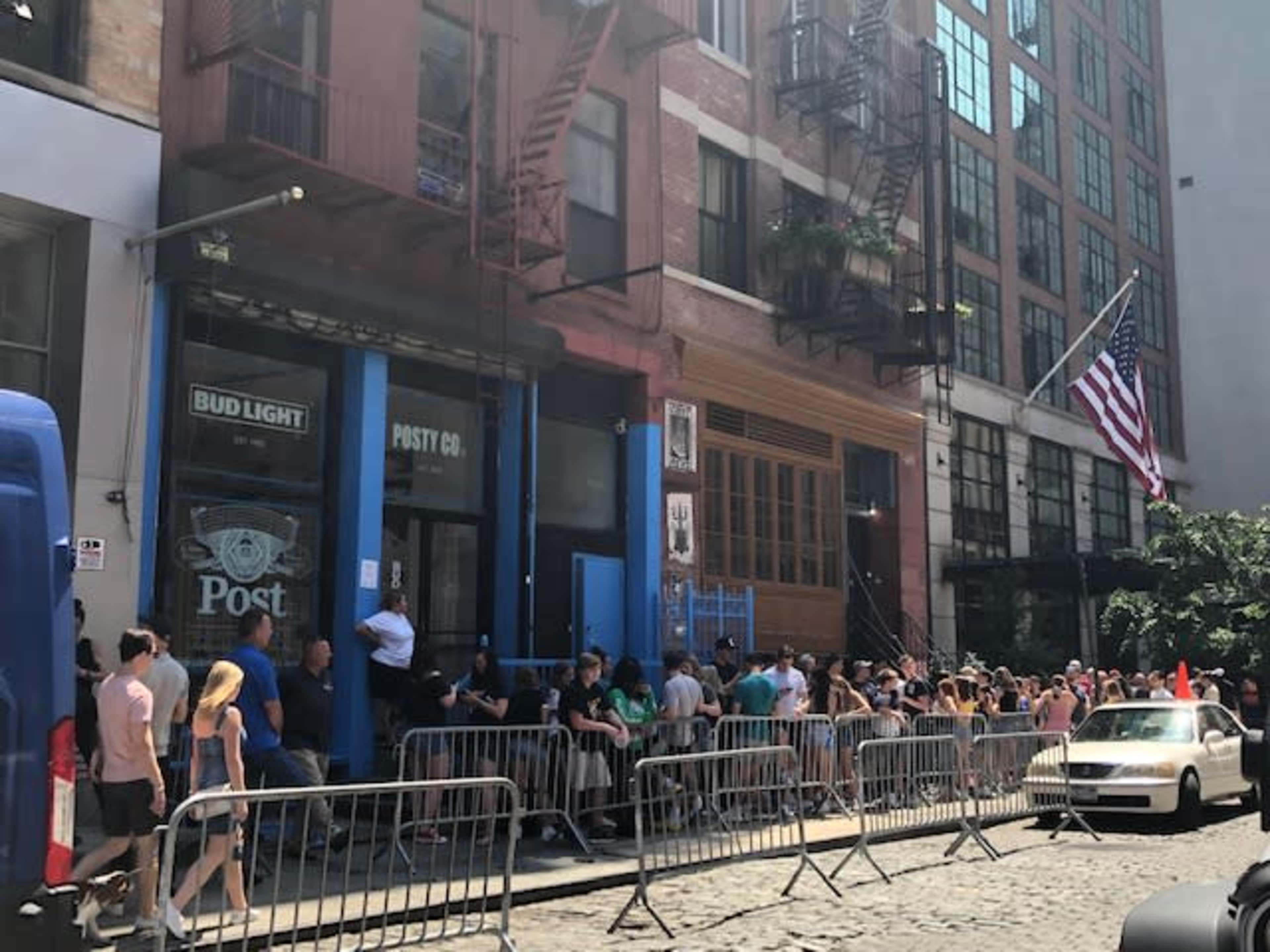 A crowd of people lines up outside a bar called "Post" on a sunny street, with an American flag visible above and barriers set up along the sidewalk.