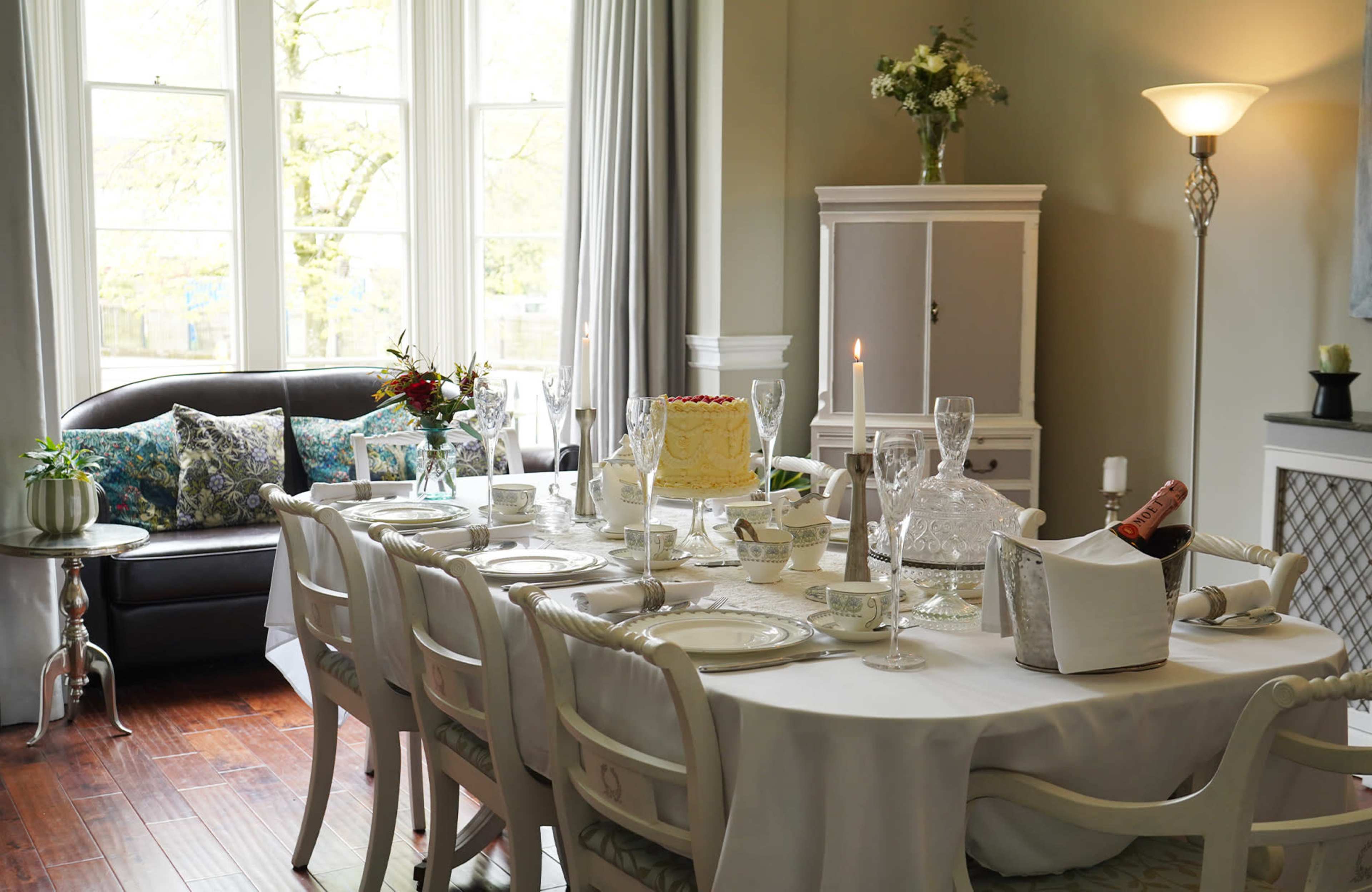 A neatly arranged dining table with white tablecloth and elegant dinnerware is set in a well-lit room featuring large windows and a comfortable sofa.