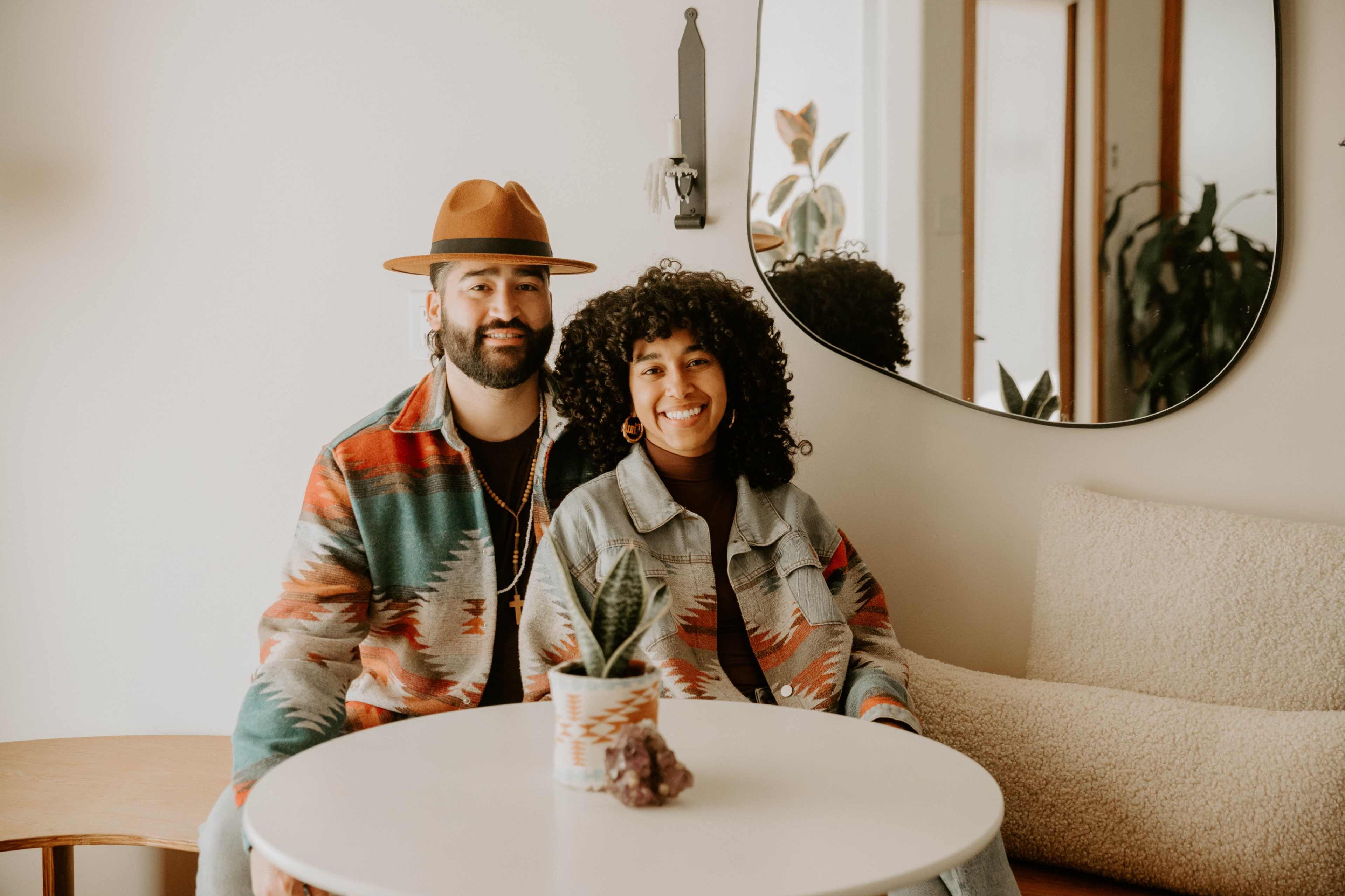 A man and a woman sit together at a small white table in a brightly lit room, surrounded by plants and decorated with a large mirror.