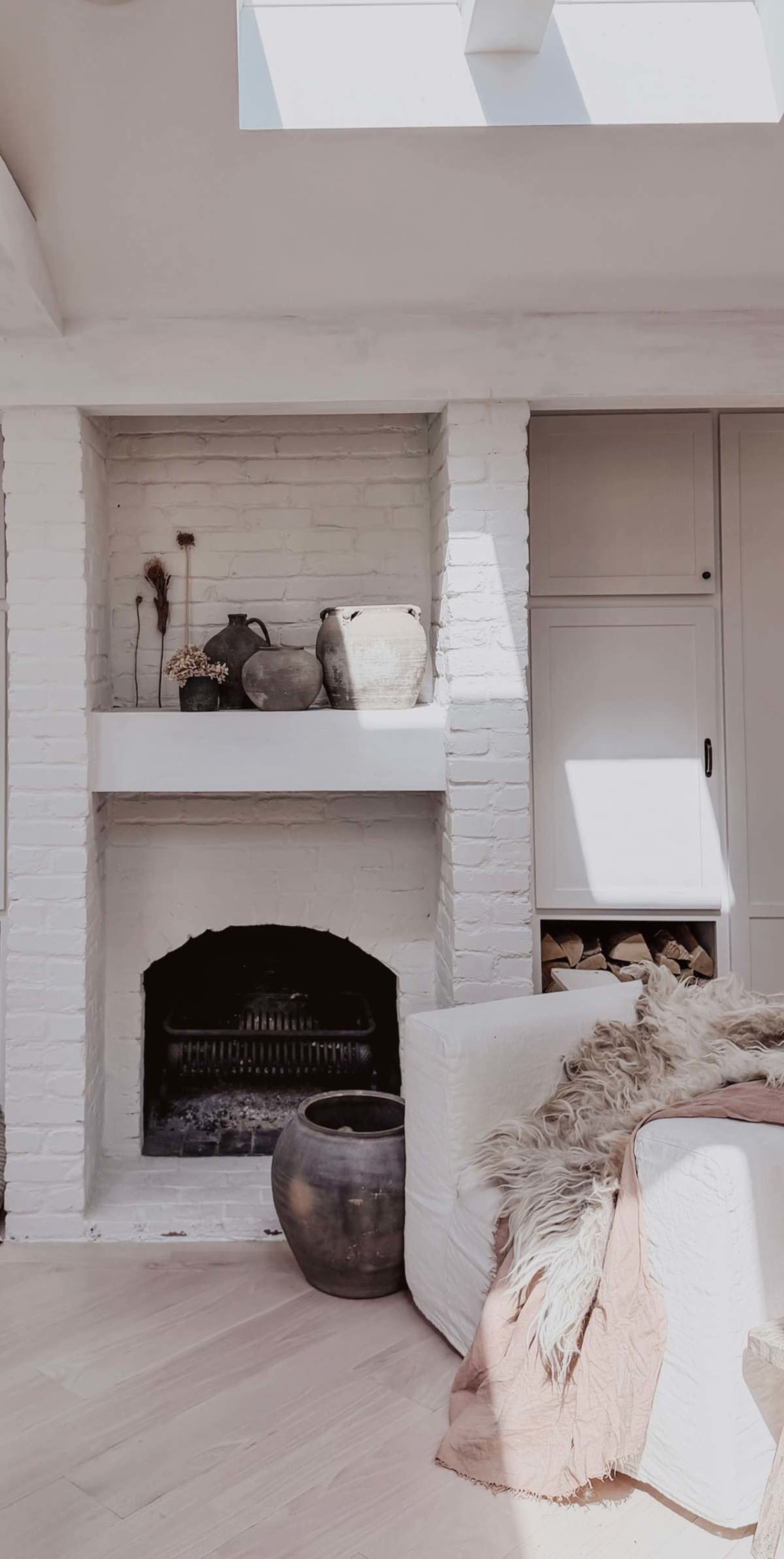 The image shows a cozy living room with a white brick fireplace, an assortment of pottery on the mantel, and a light-colored sofa adorned with a throw and a fur blanket.