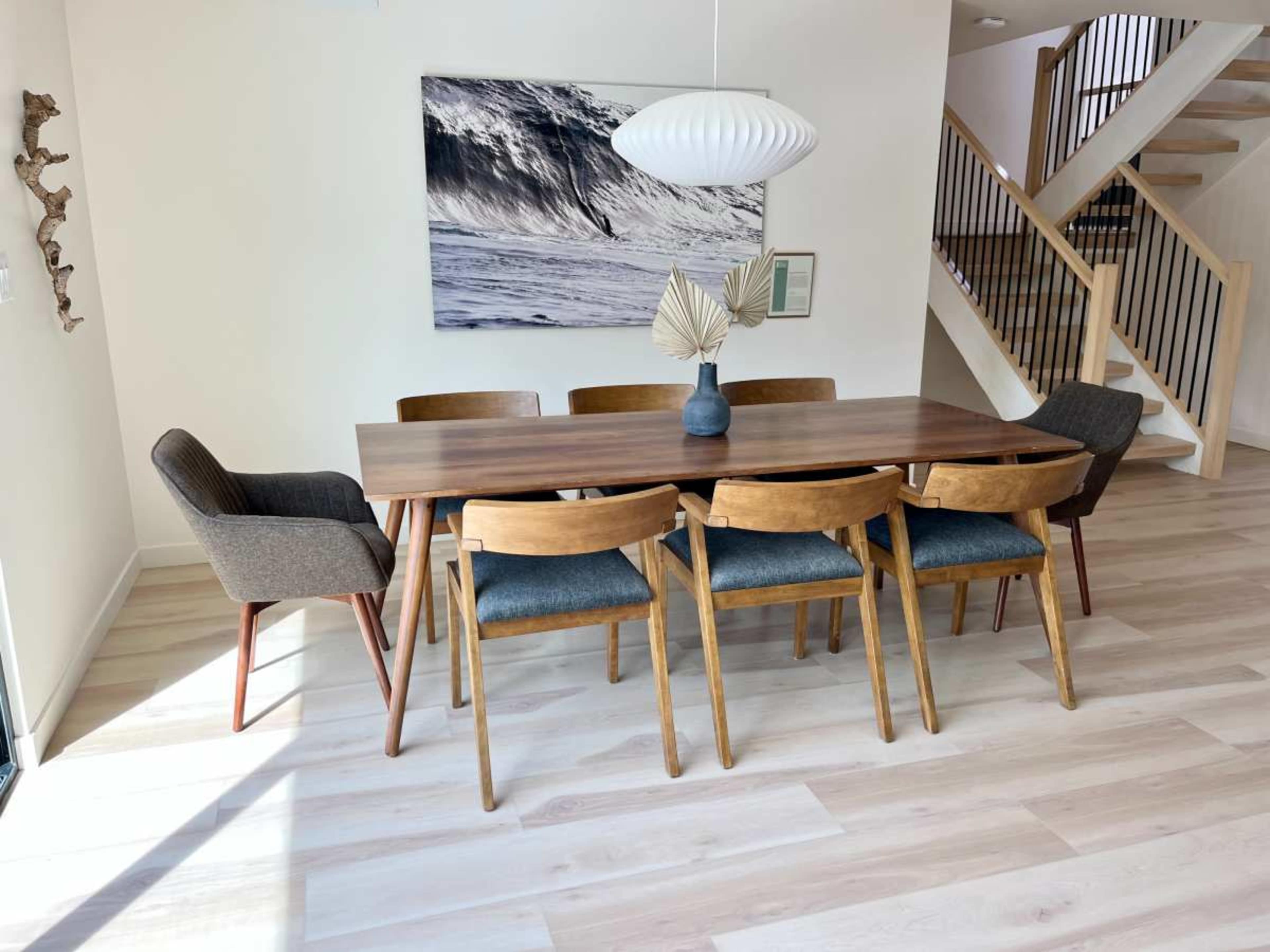 A wooden dining table with six chairs is positioned in a bright room featuring a large wave photograph on the wall and a staircase in the background.