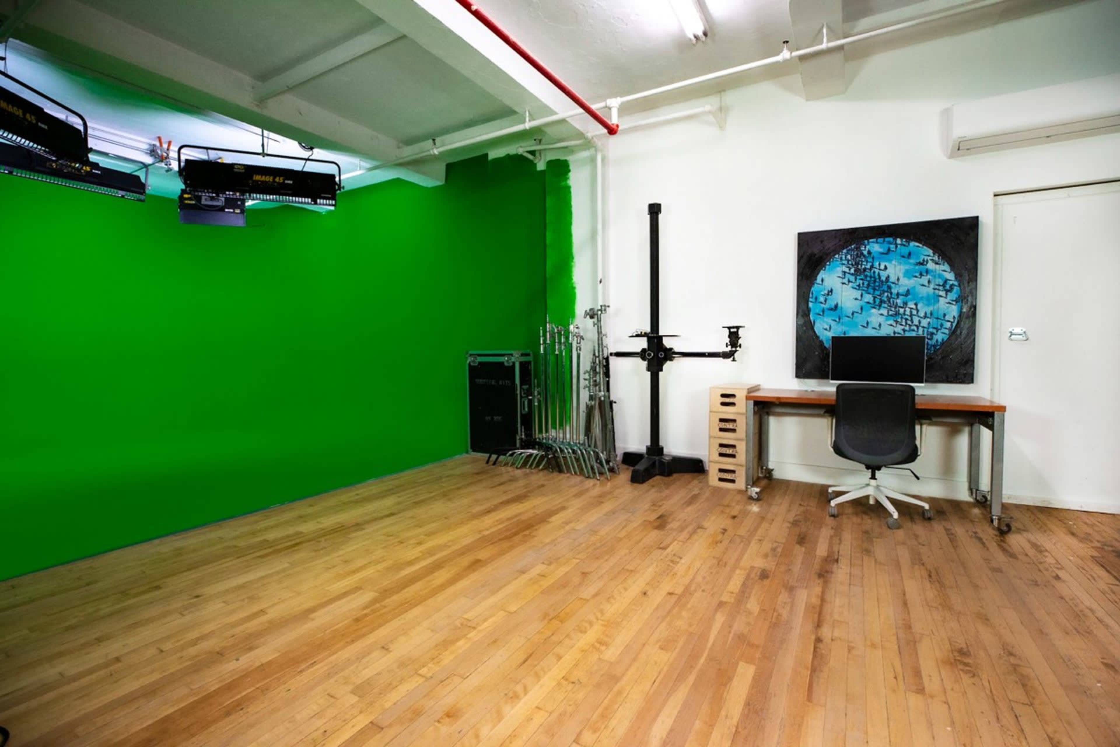 The image shows an empty studio space with a green screen on one wall, wooden flooring, and a desk with a chair.