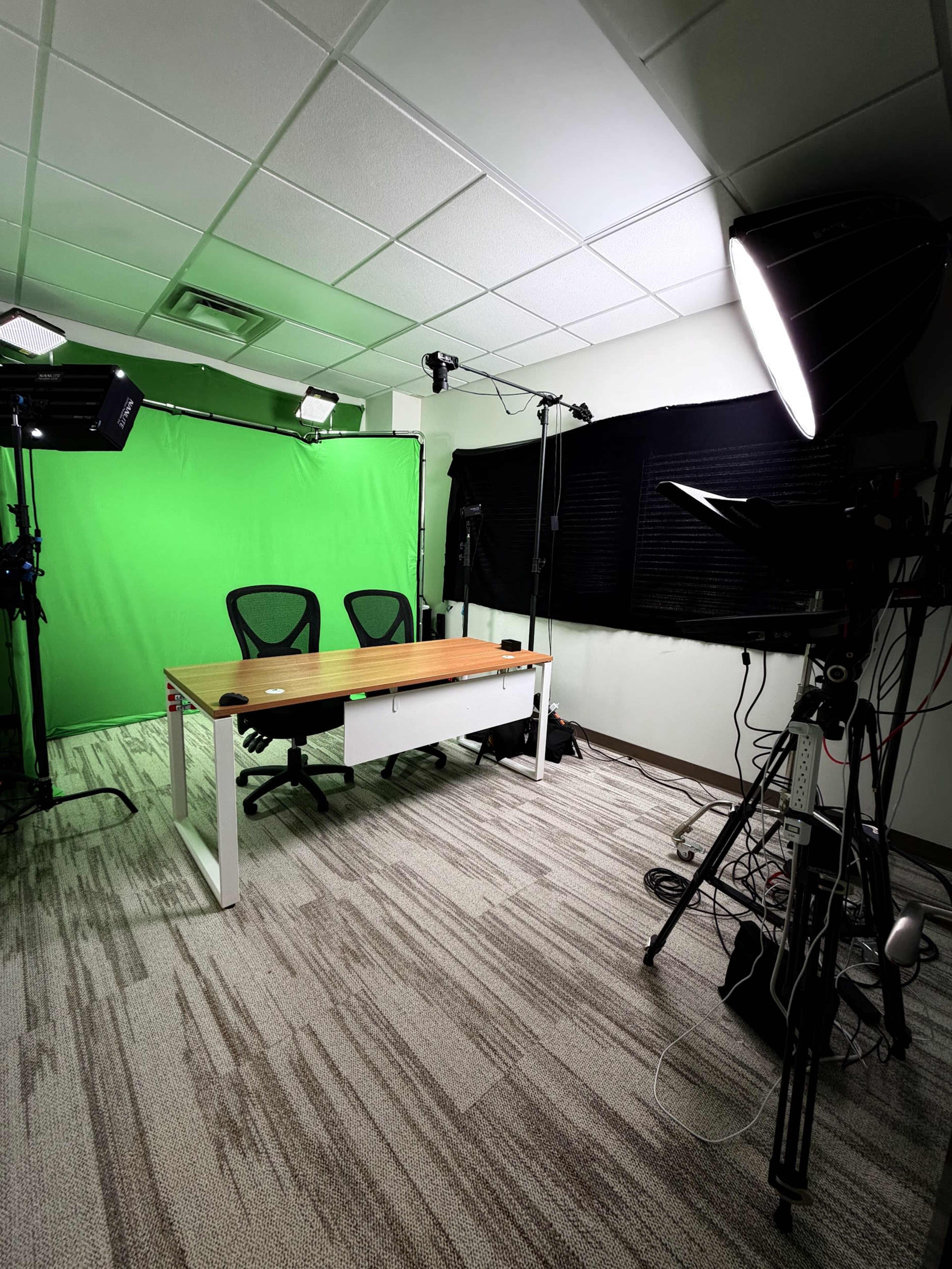 The image shows a studio space featuring two black chairs positioned in front of a wooden desk, with a green screen backdrop and various lighting equipment set up.