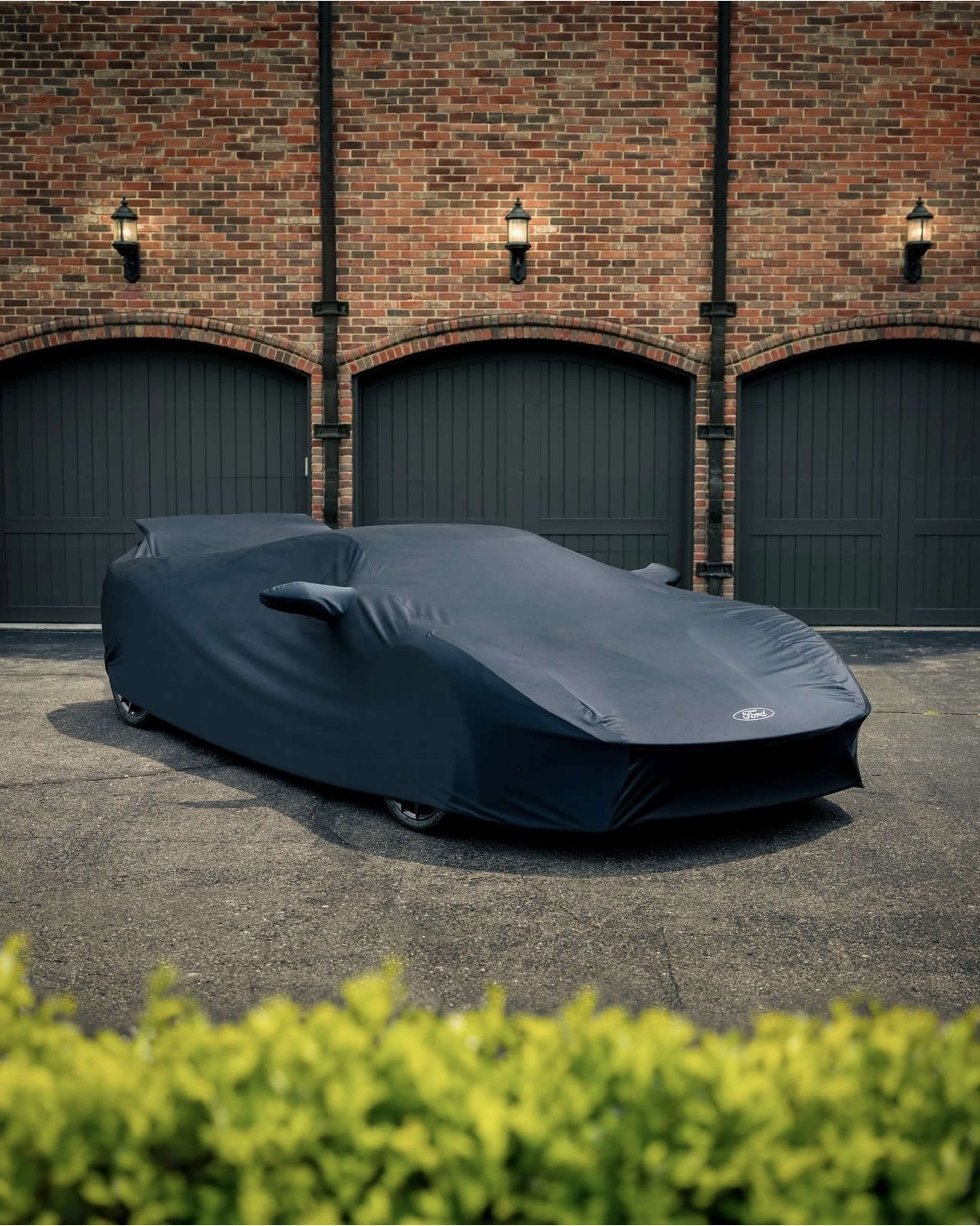 A covered sports car is parked in front of a garage with brick walls and dark wooden doors.