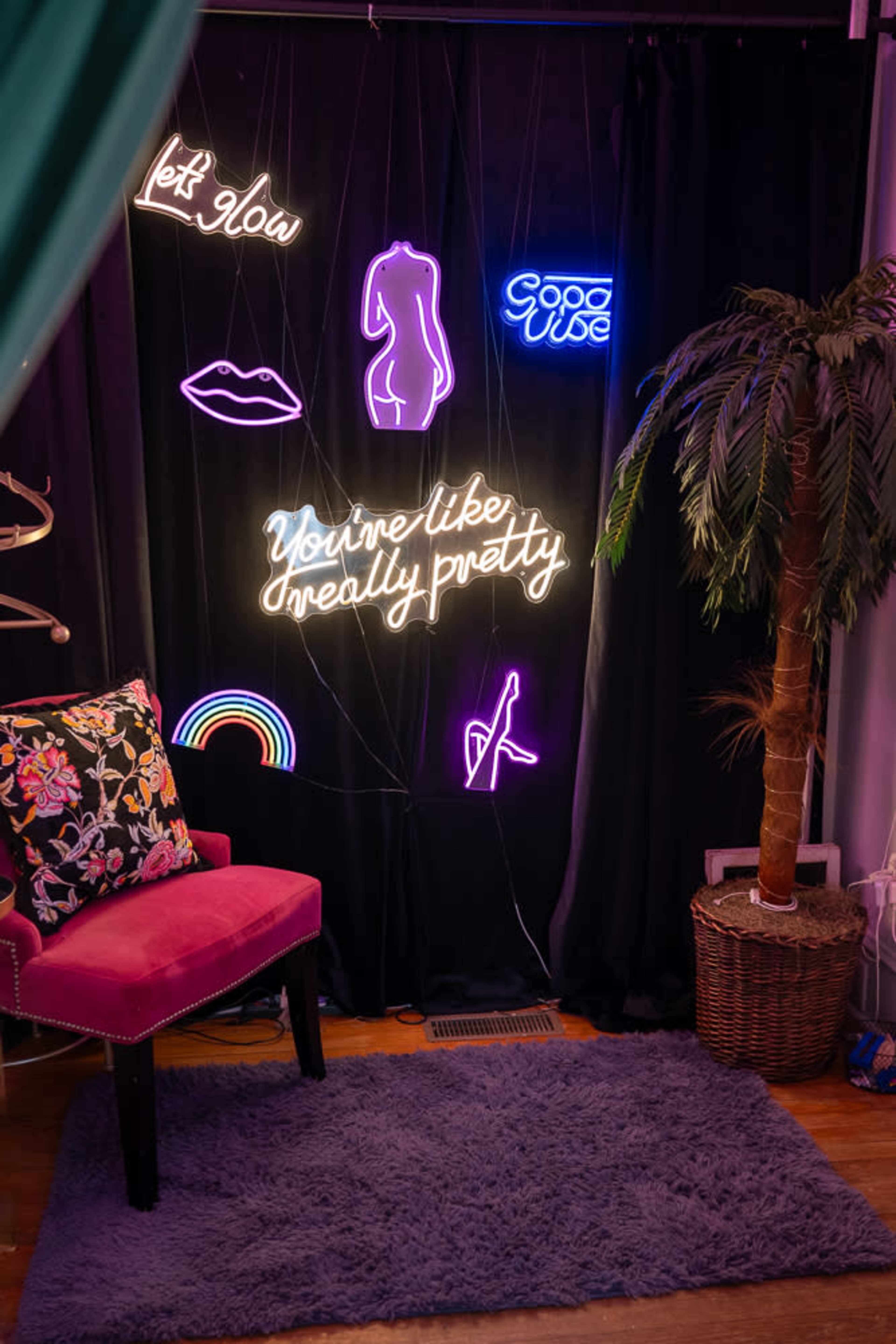 The image shows a colorful neon wall display with various lights, alongside a pink chair and a potted plant in a cozy interior setting.