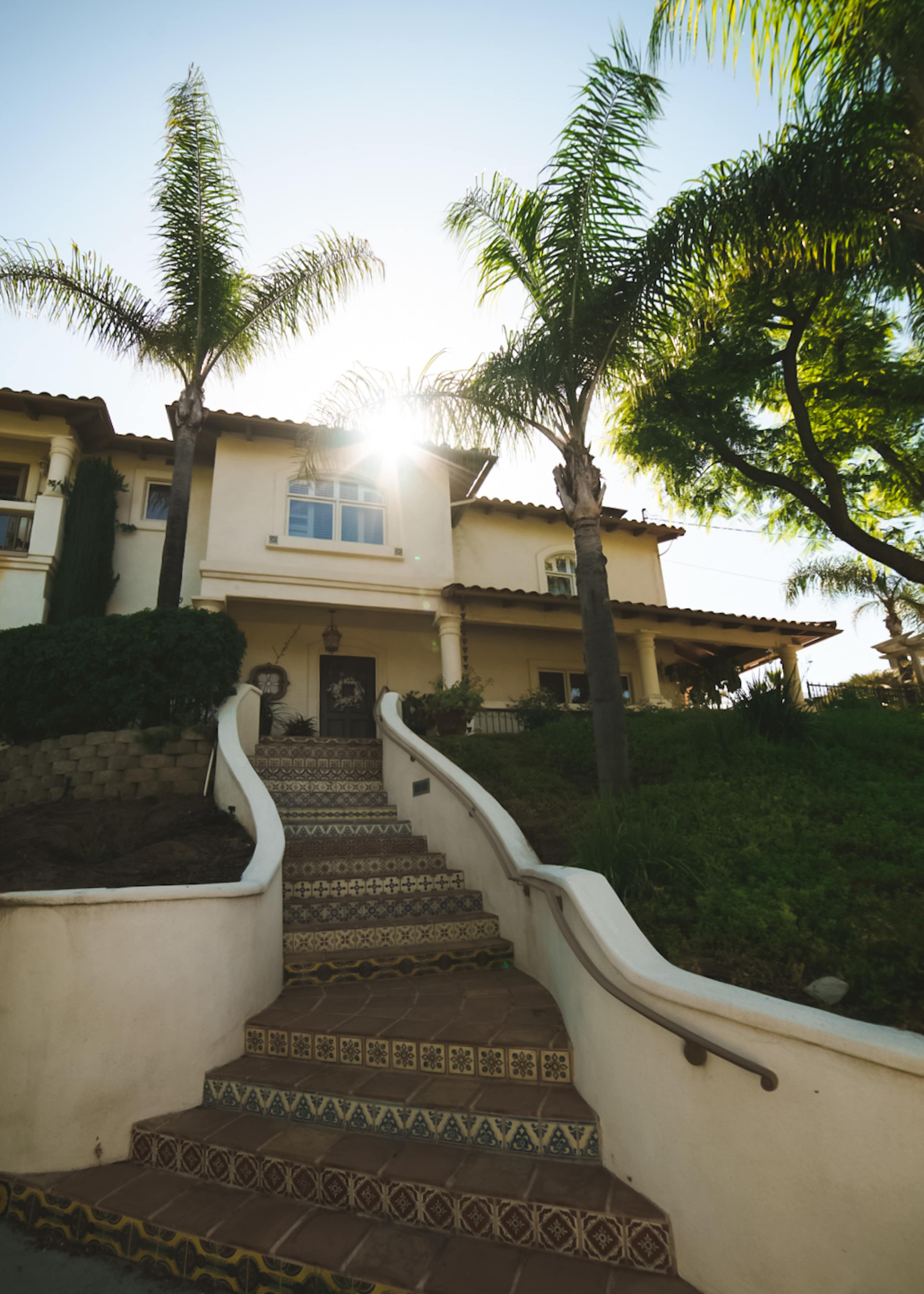 A staircase leads up to a two-story house adorned with palm trees and sunlight shining in the background.
