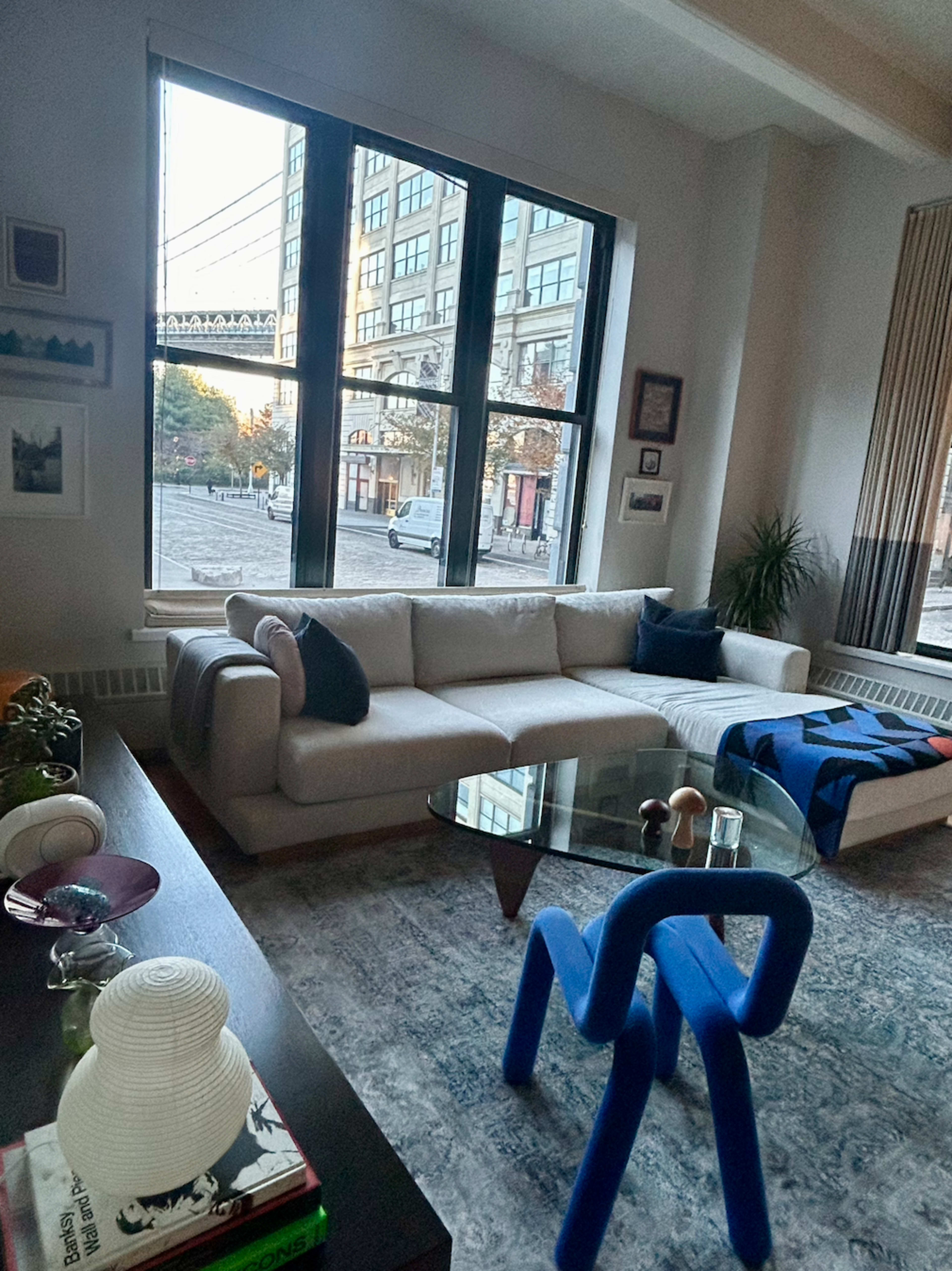The interior of a living room features a light-colored sofa, a glass coffee table, and large windows overlooking a city street.