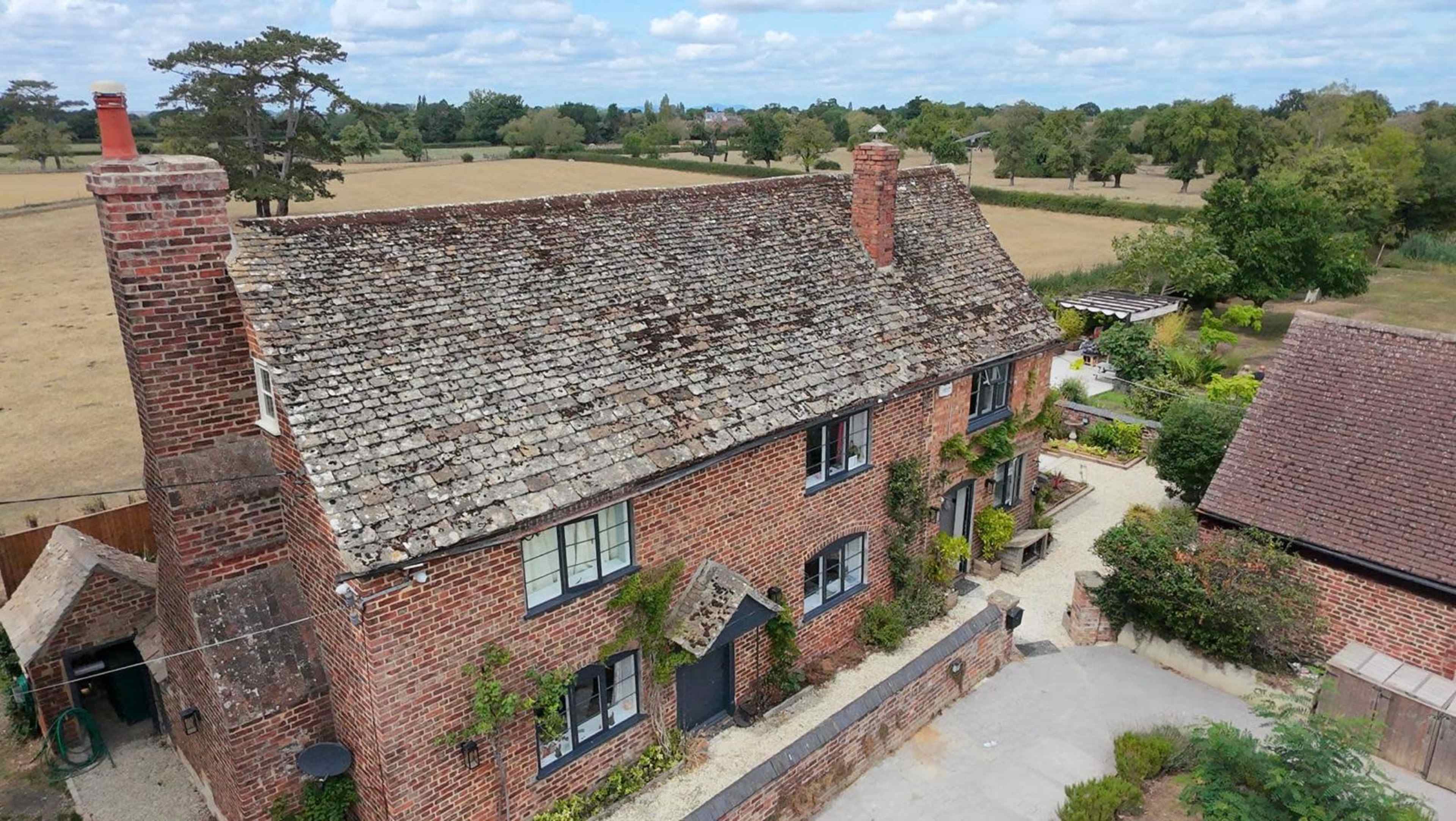 The image shows a rustic brick house with a sloped roof, surrounded by greenery and open fields, under a partly cloudy sky.