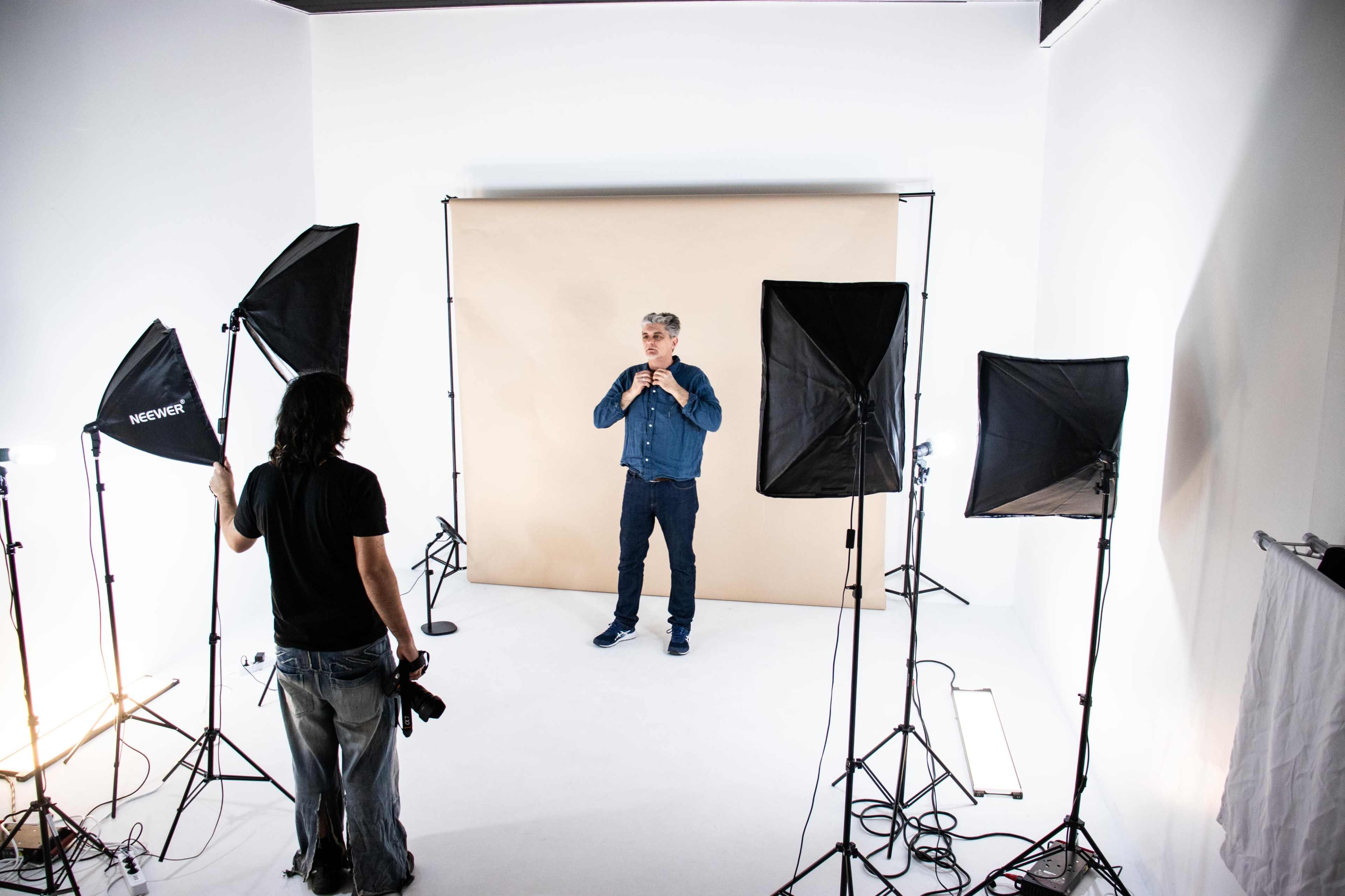 A photographer is preparing to take a portrait of a man standing in front of a beige backdrop in a studio lit by several softboxes.