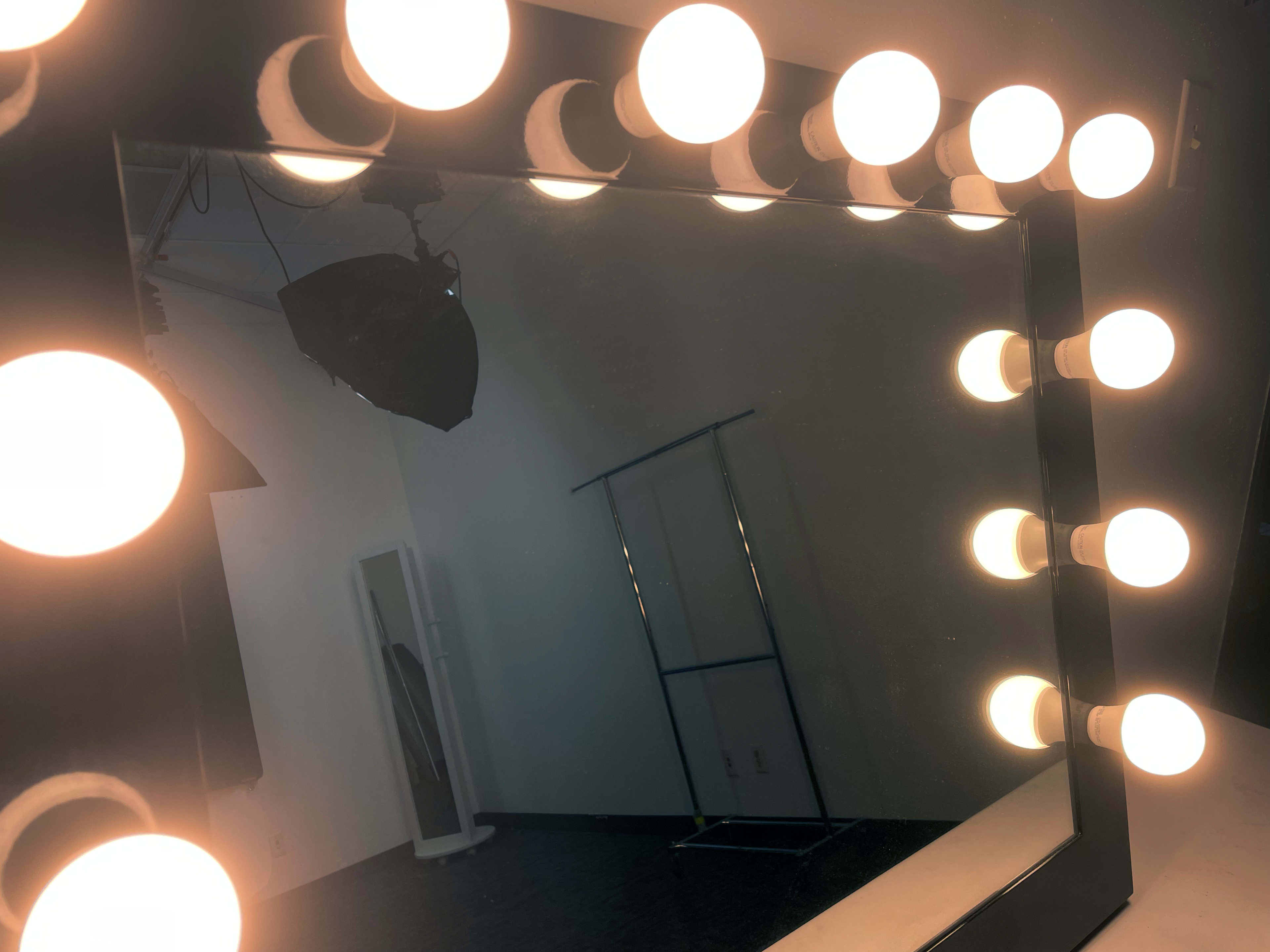 A large vanity mirror is surrounded by light bulbs, reflecting a dimly lit room with a clothing rack in the background.