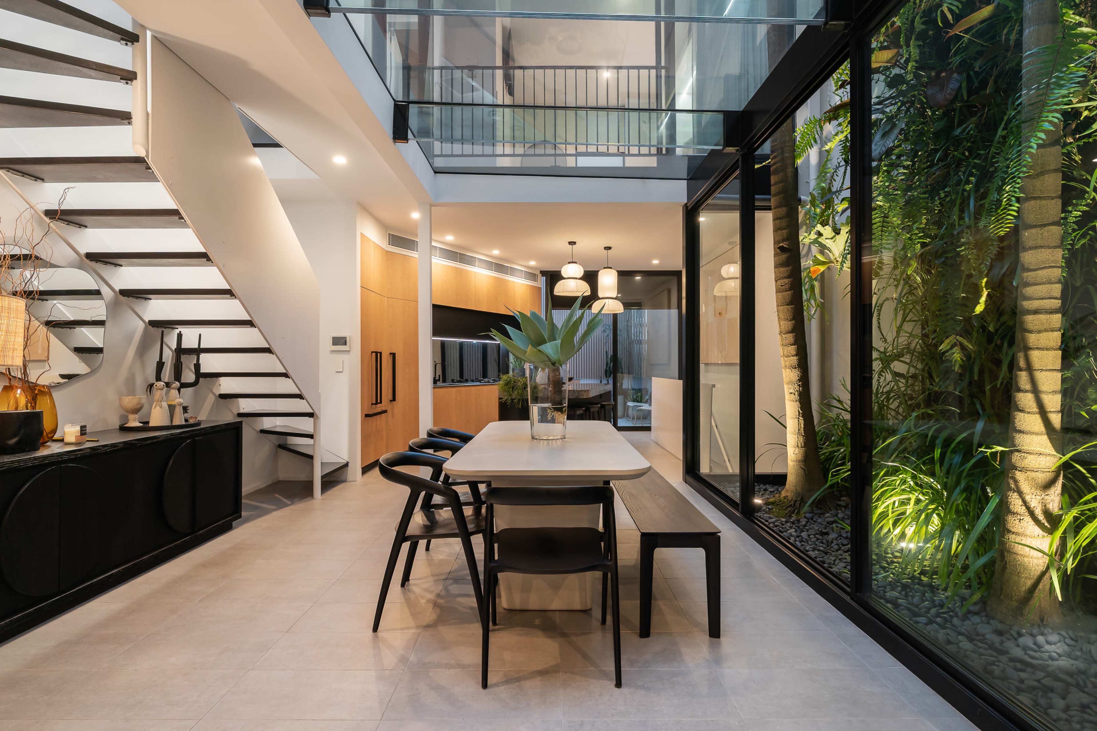 The image shows a modern dining area with a long table and black chairs, surrounded by large windows and indoor plants, adjacent to a staircase.